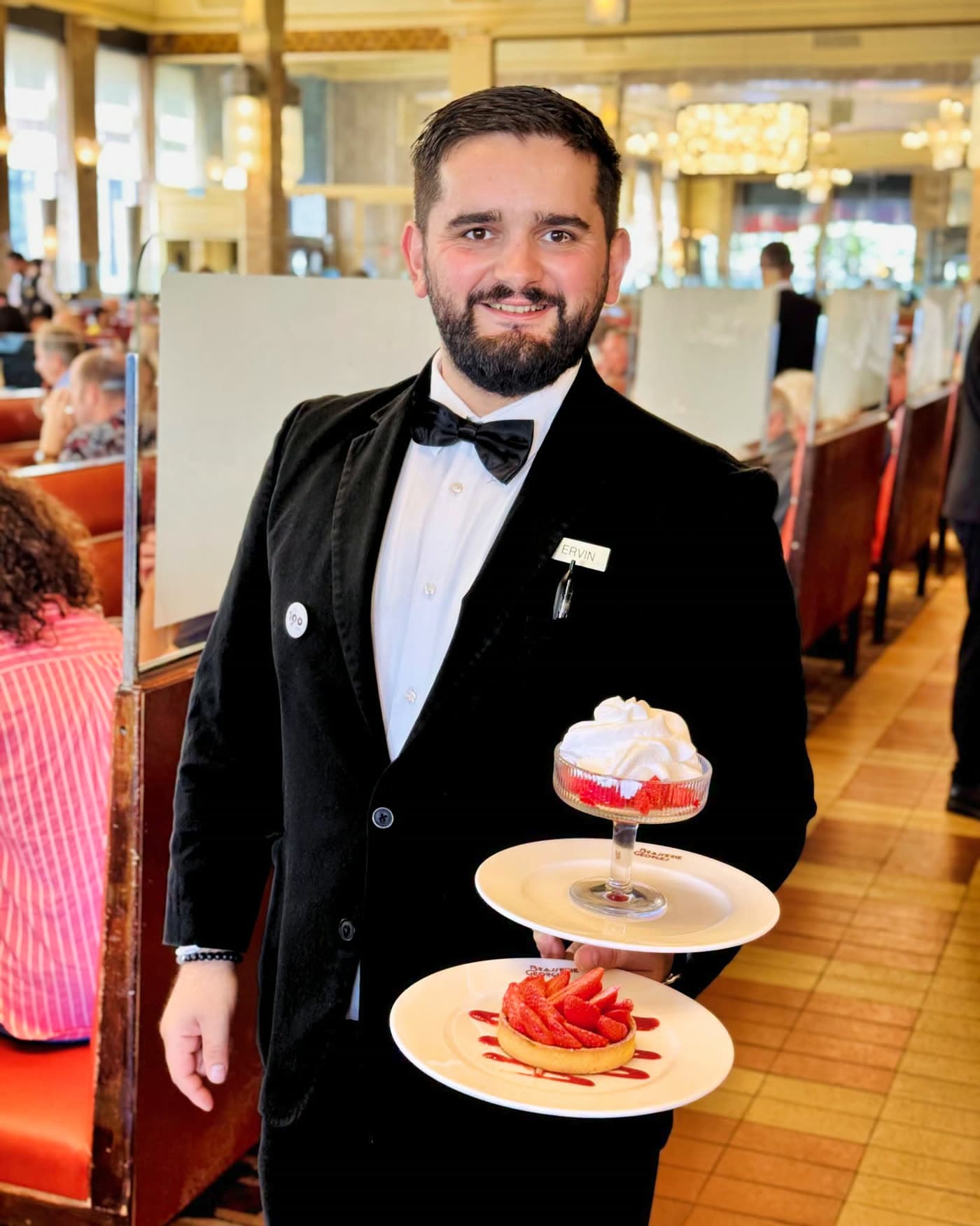 Serveur en smoking souriant tenant une coupe de fraises chantilly et une tarte aux fraises — Brasserie Georges