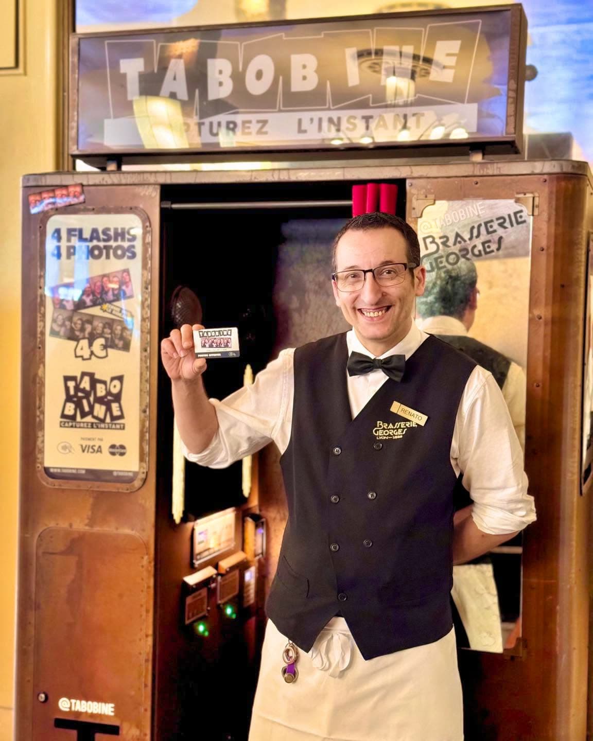 Serveur souriant devant une cabine photomaton vintage, tenant une bande de photos — Brasserie Georges