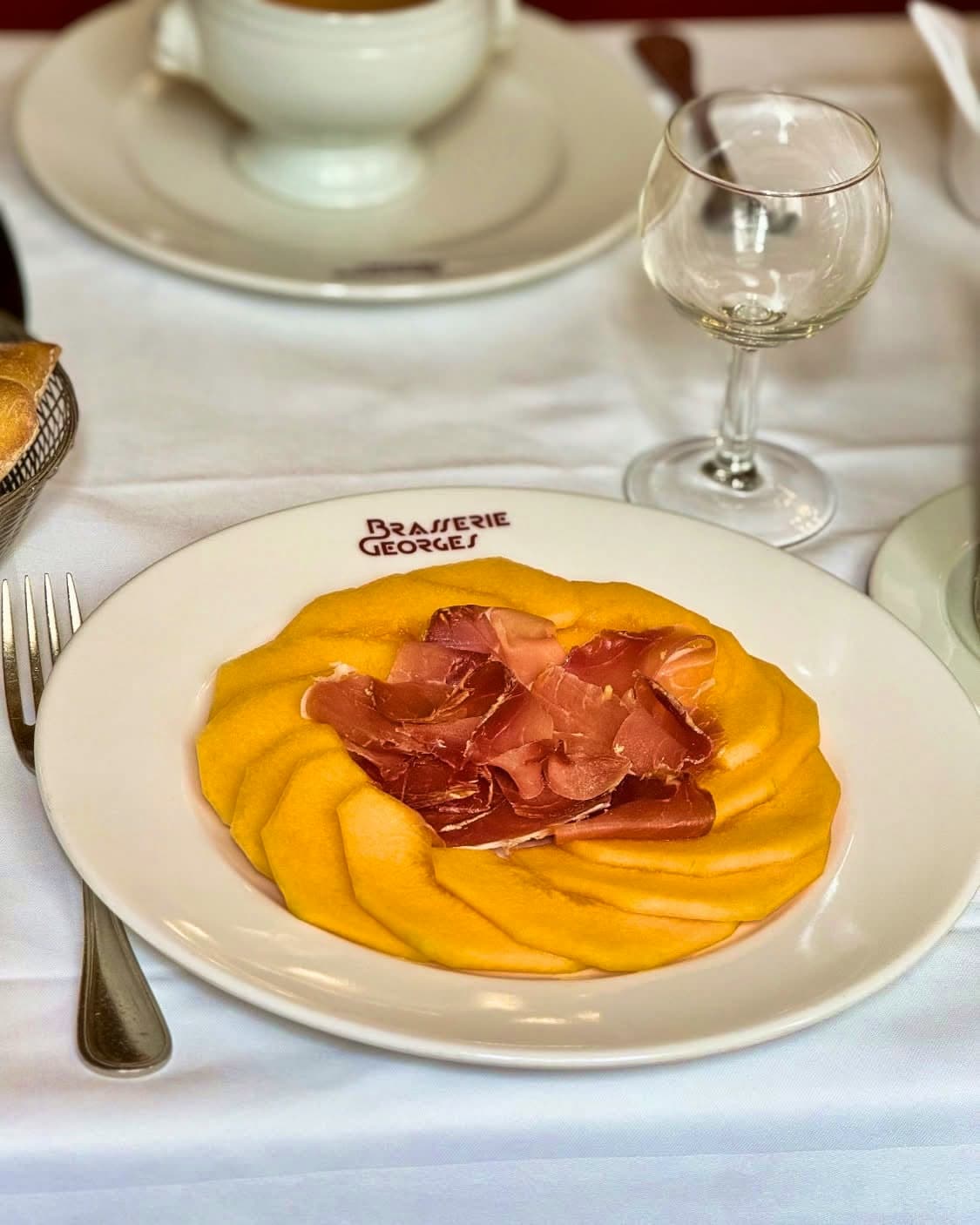 Assiette de melon et jambon cru disposés en rosace, verre à vin et pain sur nappe blanche — Brasserie Georges