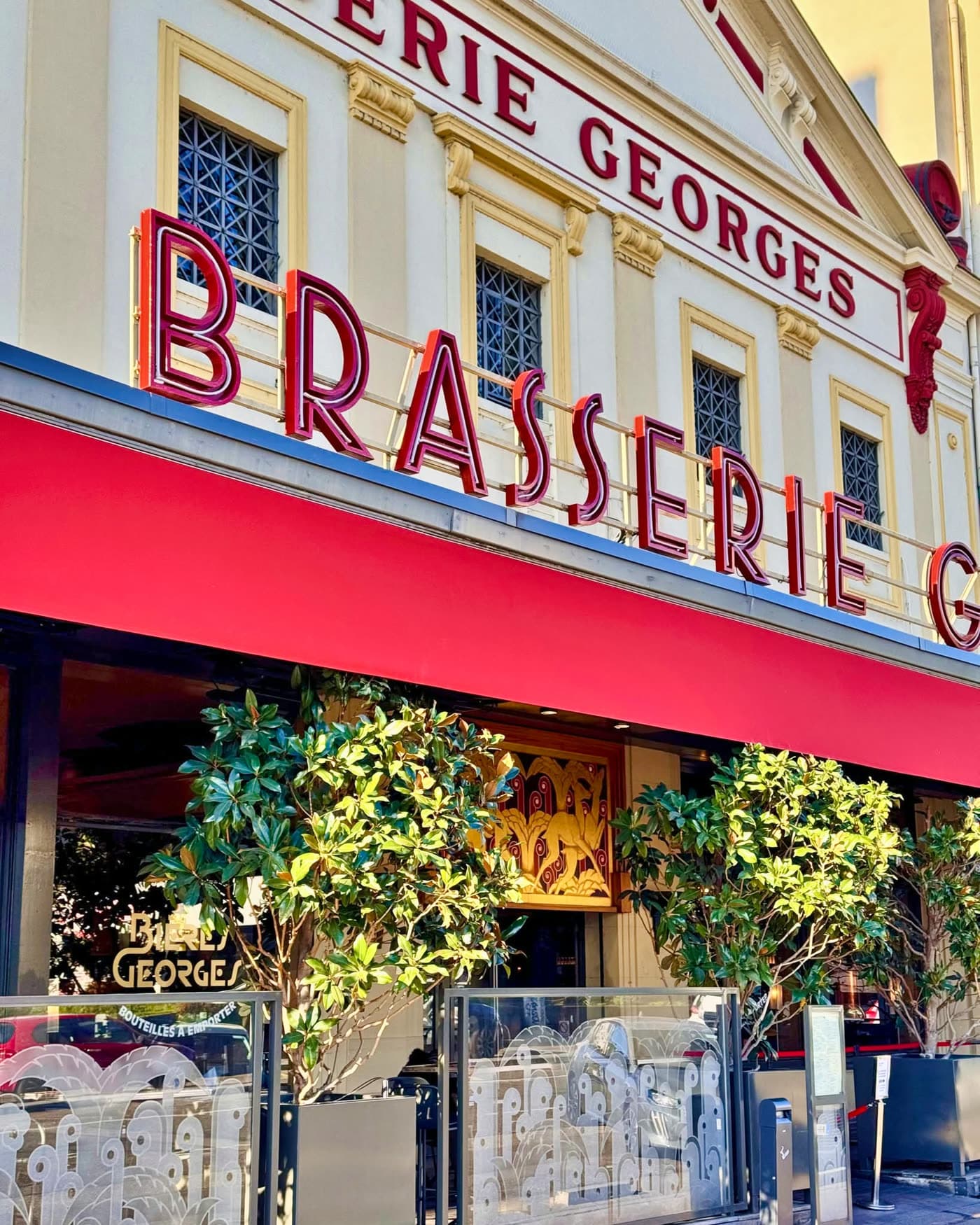 Cadrage serré sur enseigne BRASSERIE GEORGES rouge, auvent, plantes en pot