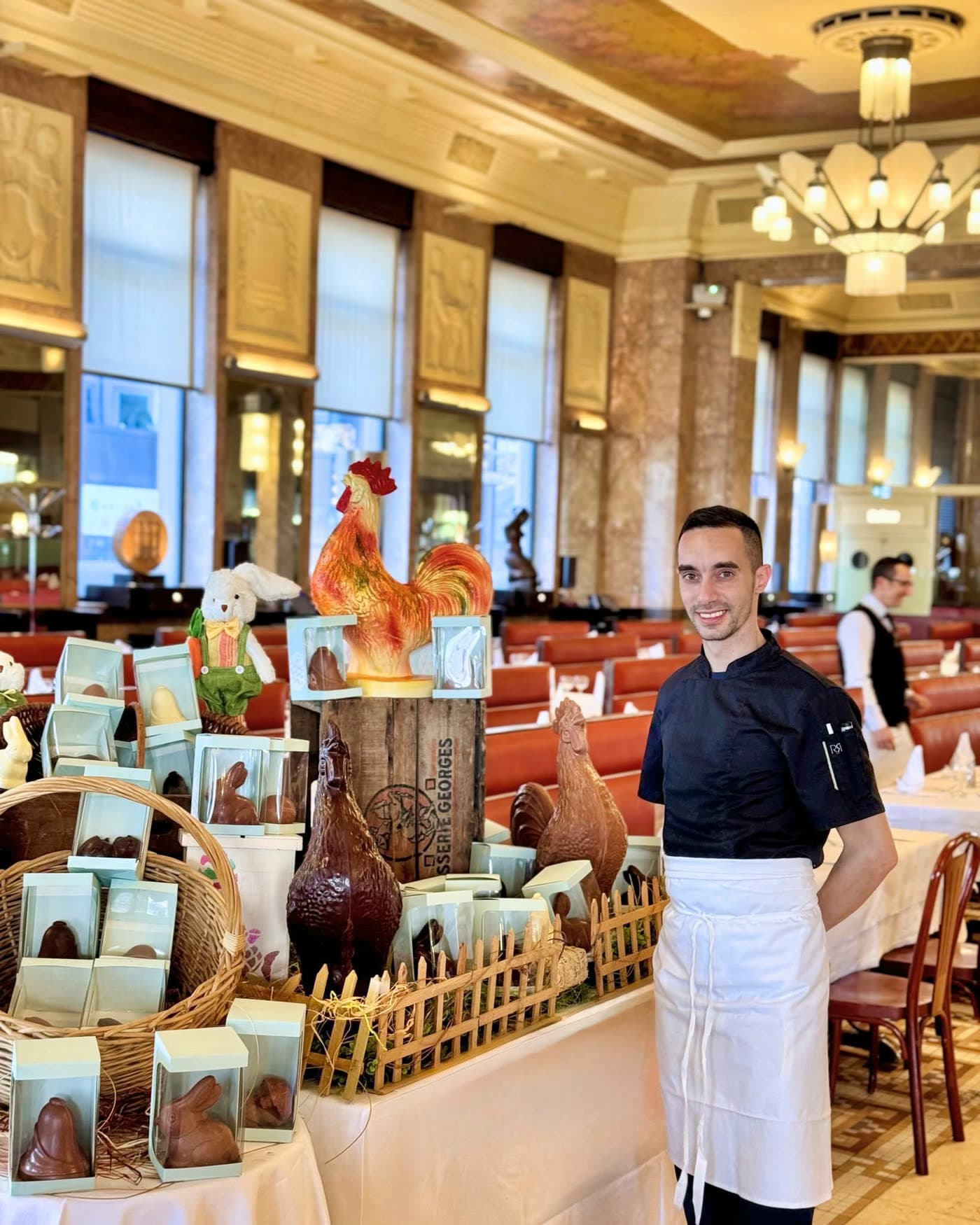Employé souriant devant présentoir de chocolats de Pâques (poules, lapins) — Brasserie Georges