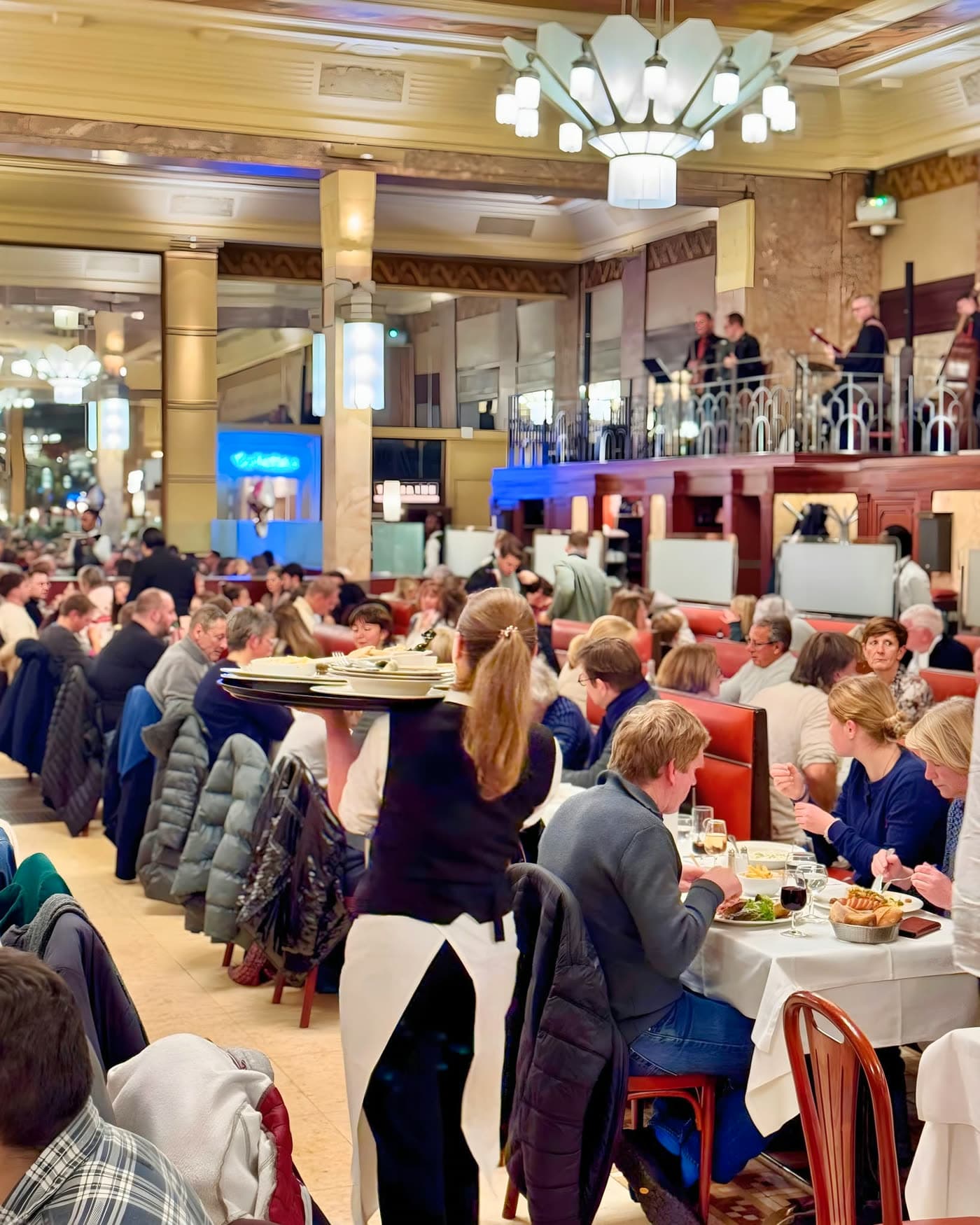 Serveuse portant une assiette traverse la salle bondée, mezzanine avec musiciens en fond — Brasserie Georges