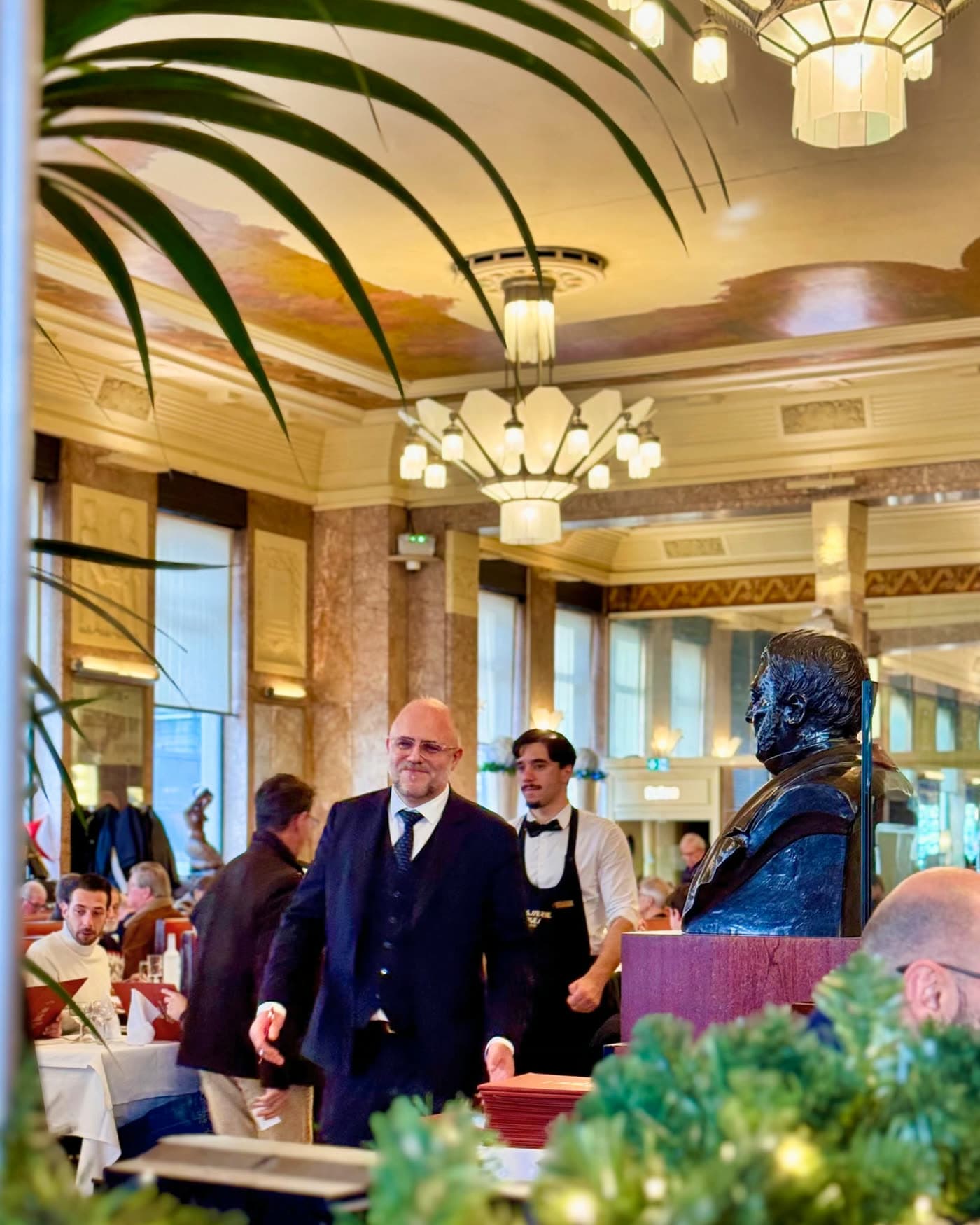 Maître d'hôtel en costume traverse la salle, buste du fondateur et feuillage au premier plan — Brasserie Georges