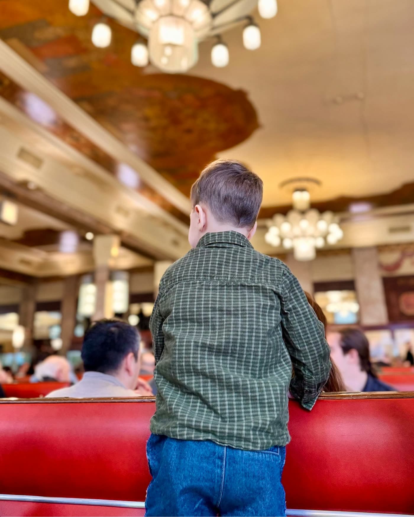 Jeune enfant de dos debout sur une banquette, regarde le plafond peint et les lustres — Brasserie Georges
