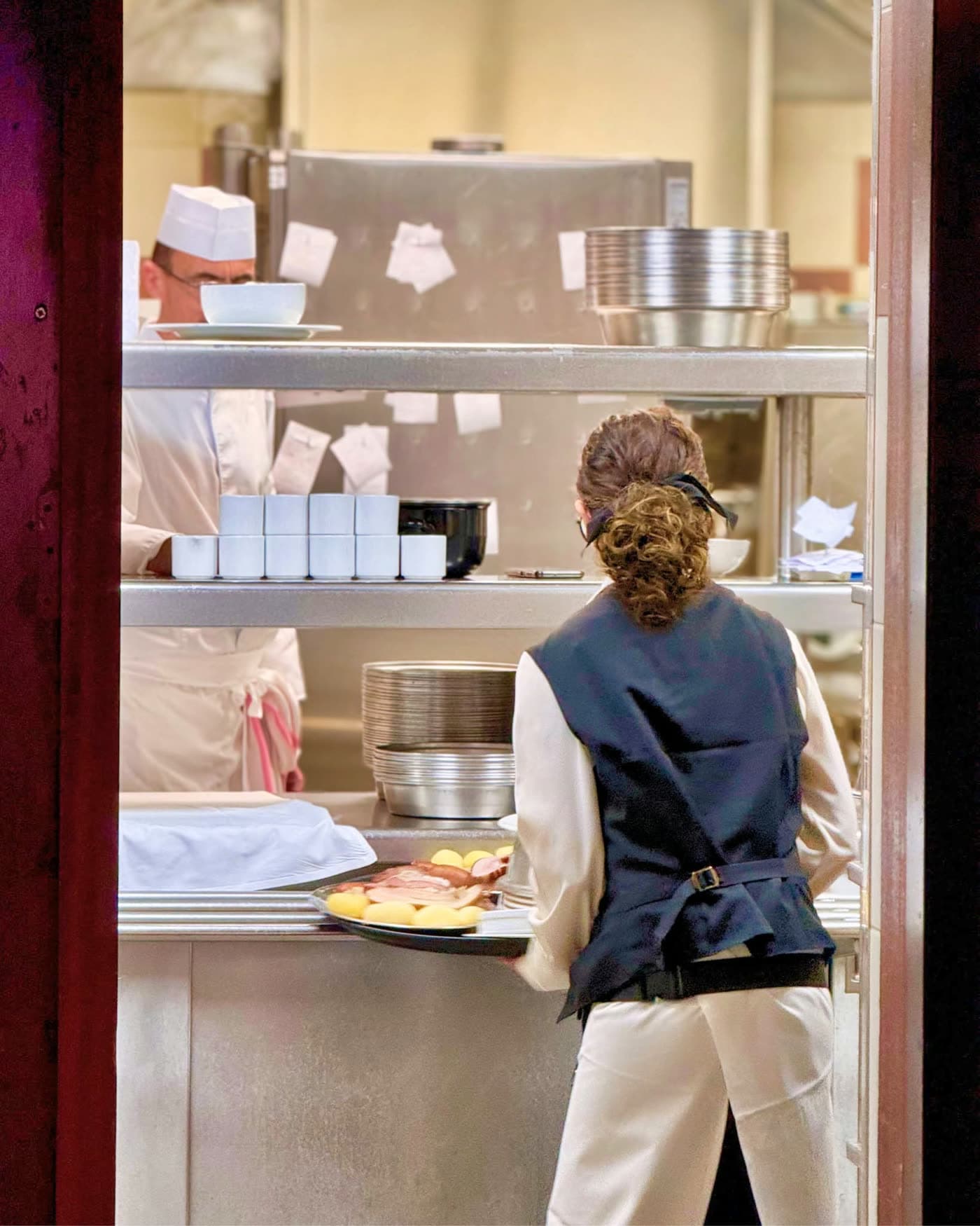 Serveuse de dos au passe avec cuisinier en toque en arrière-plan — Brasserie Georges