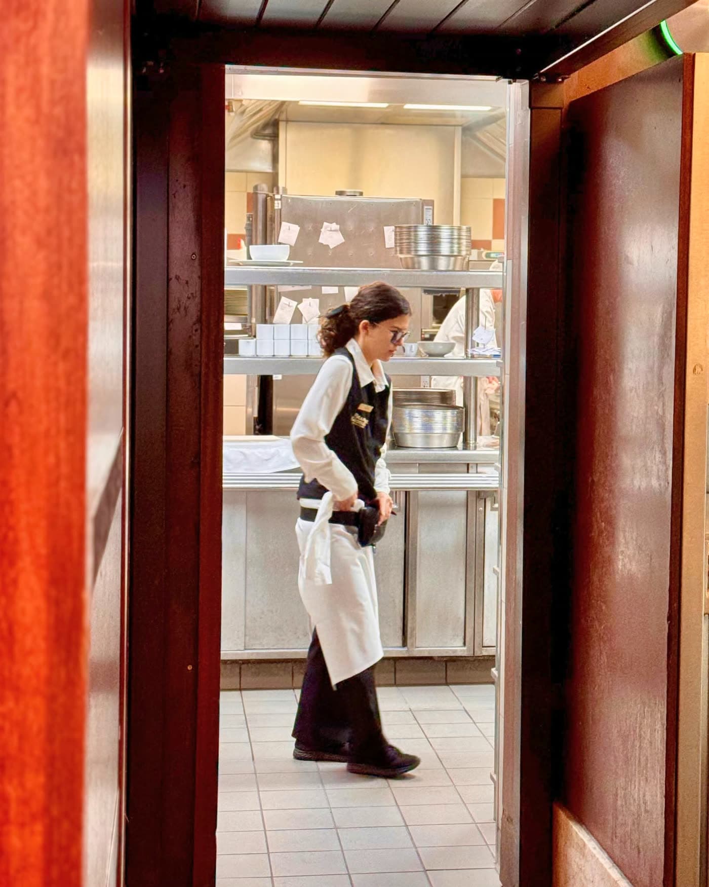 Employée en tablier blanc traversant une porte en bois acajou vers la cuisine — Brasserie Georges