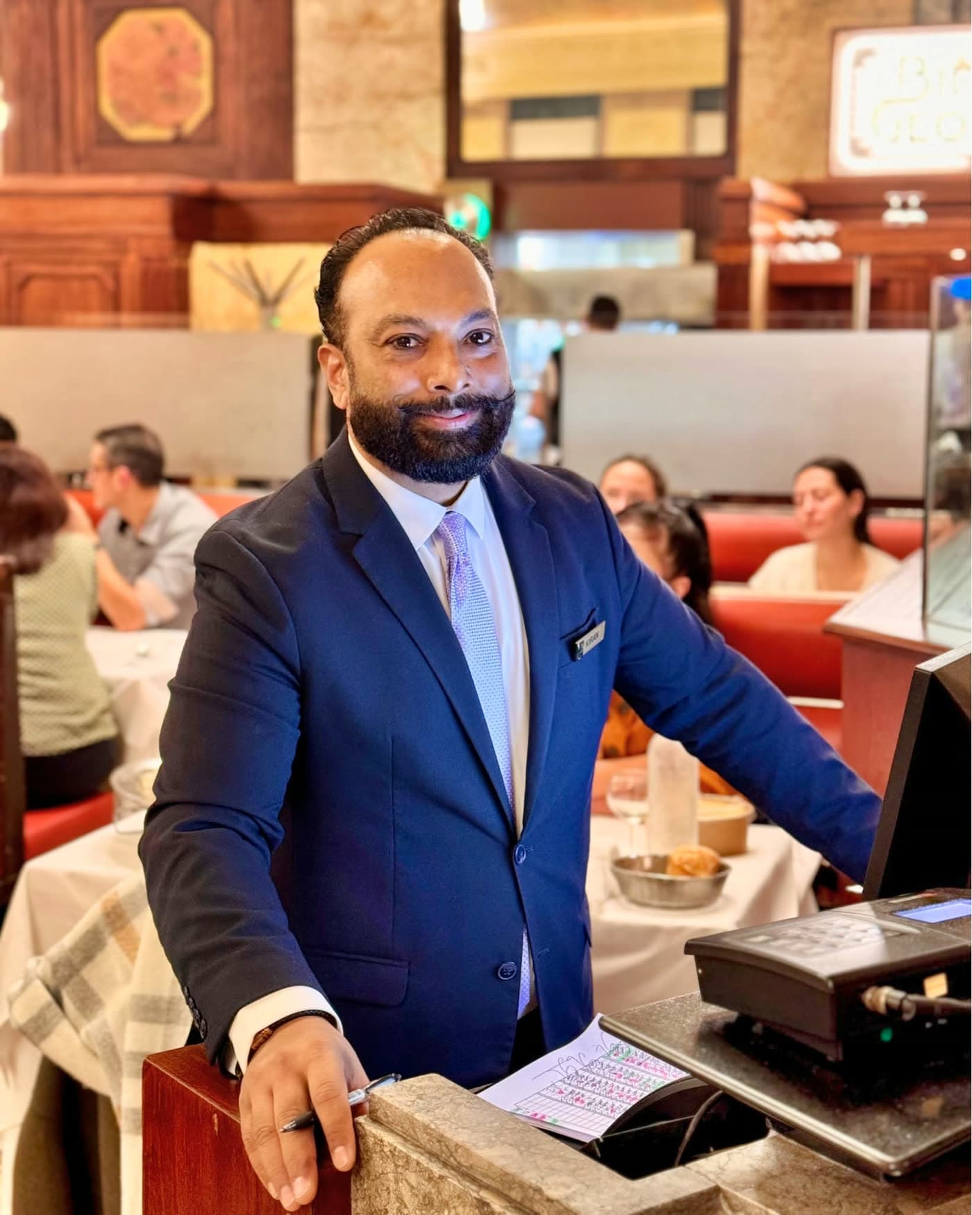 Maître d'hôtel en costume bleu marine souriant au comptoir d'accueil, salle en fond — Brasserie Georges