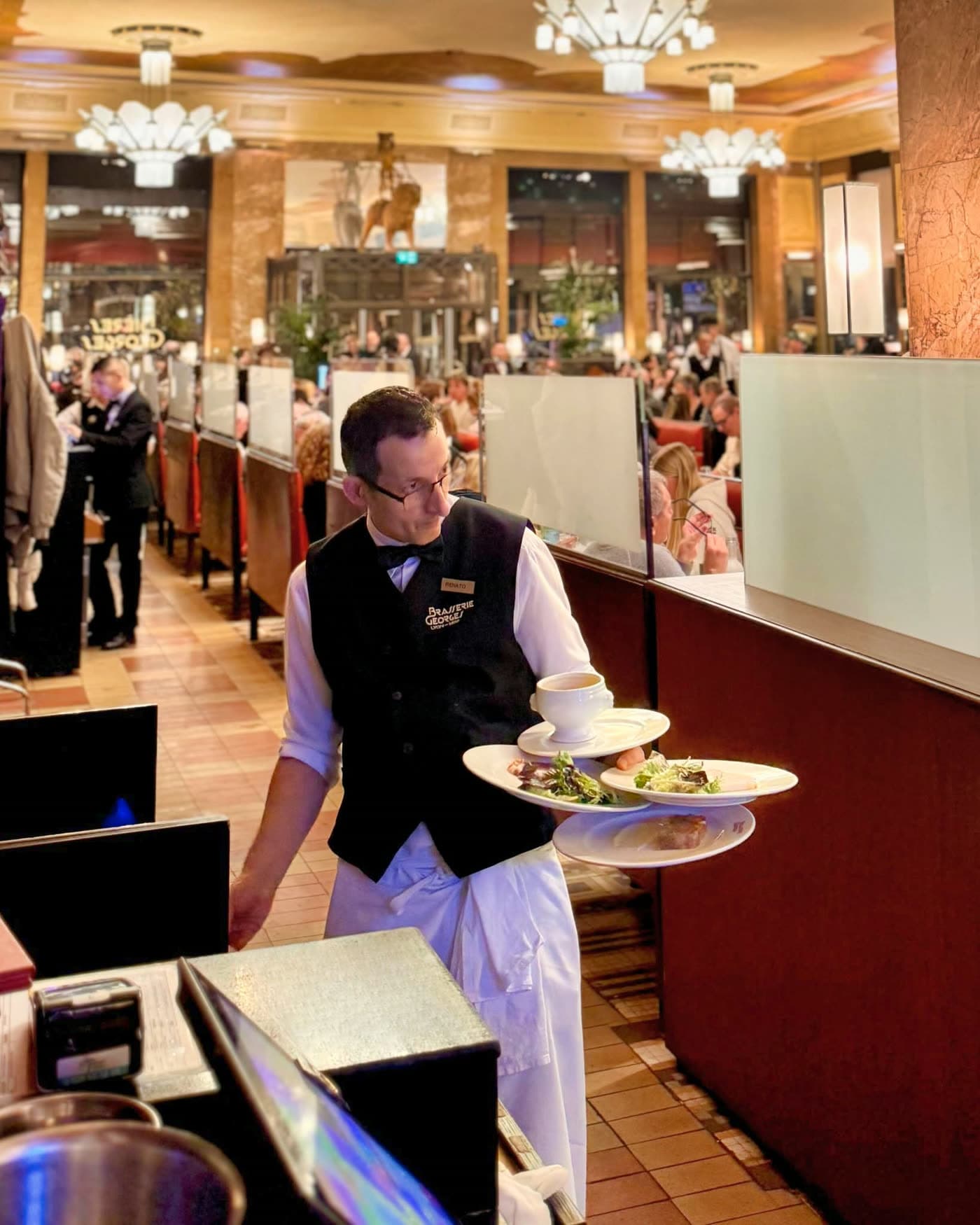 Serveur portant plusieurs assiettes garnies en traversant la salle, clients flous en fond — Brasserie Georges