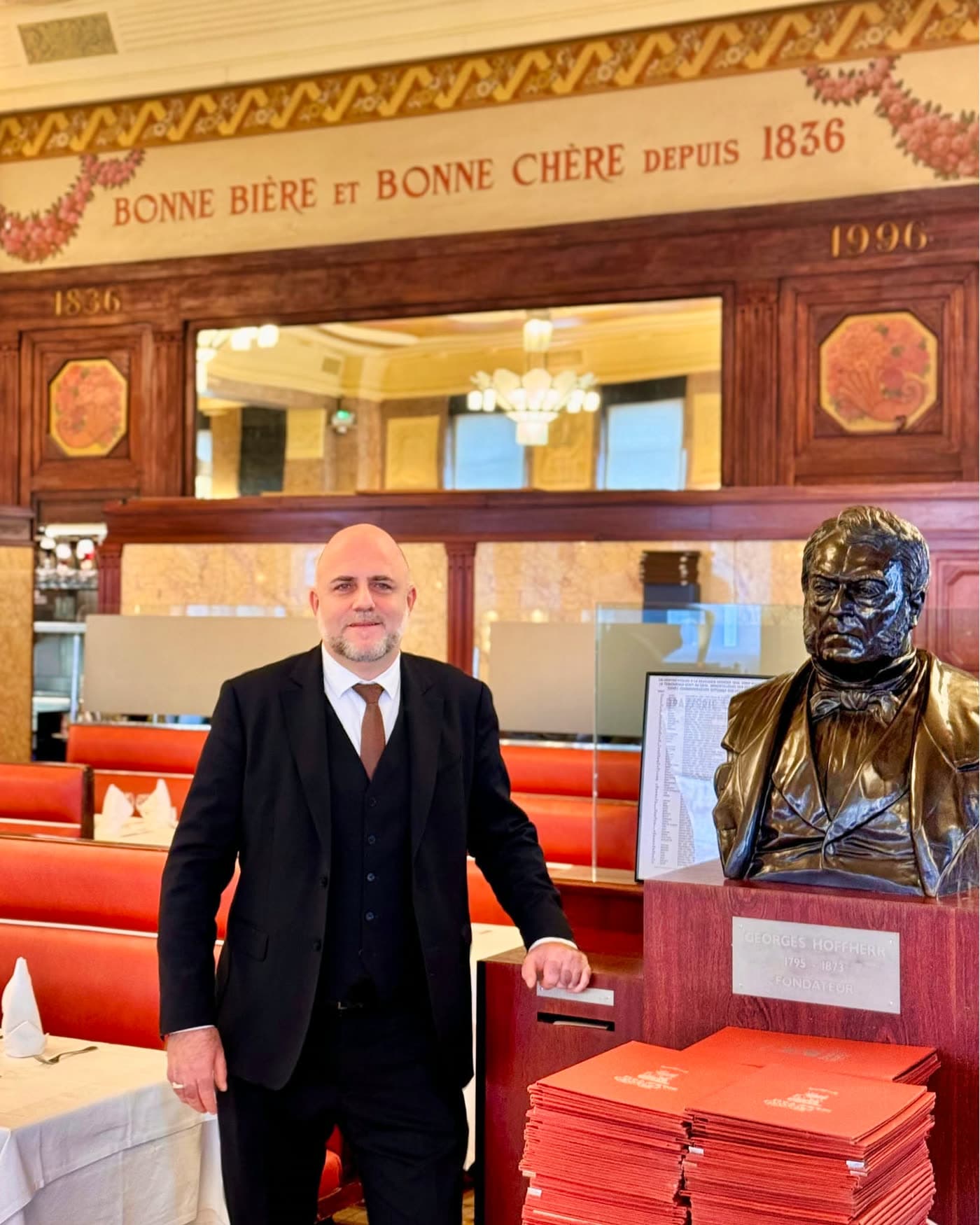 Homme en costume posant près du buste du fondateur sous fresque historique — Brasserie Georges