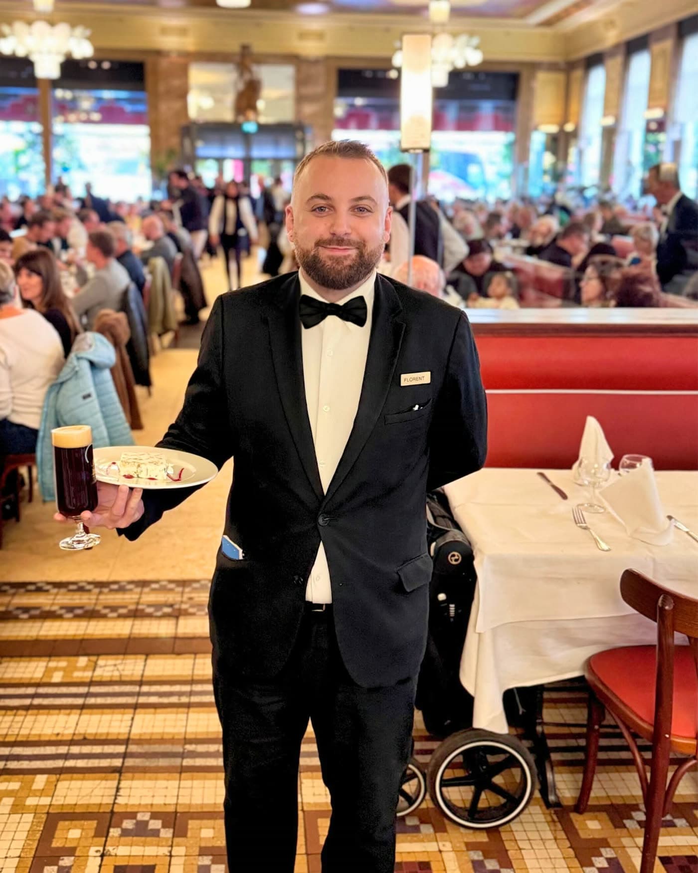 Maître d'hôtel en smoking portant une assiette et une bière, salle pleine en fond — Brasserie Georges