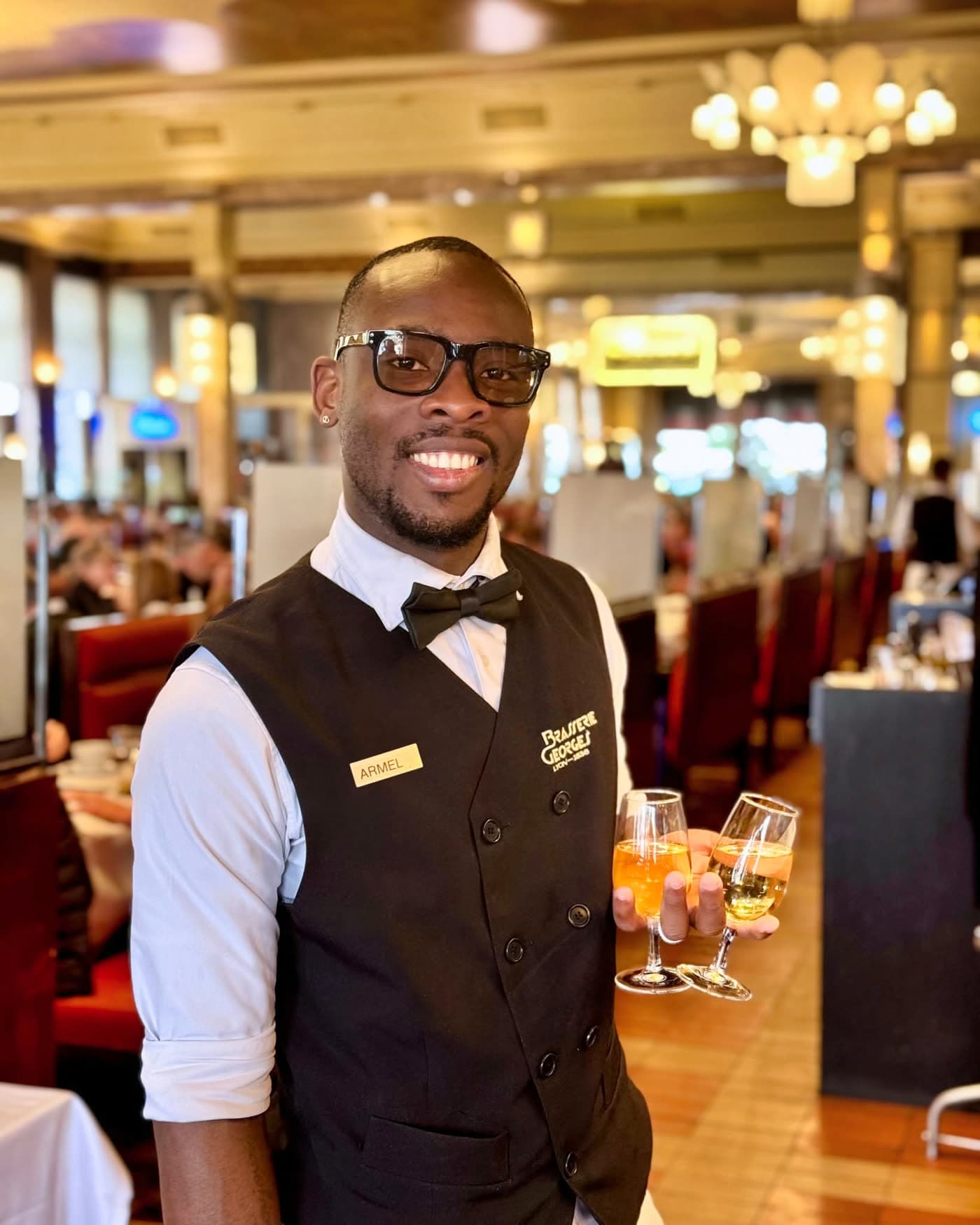 Portrait souriant d'un serveur en gilet et noeud papillon tenant deux cocktails — Brasserie Georges