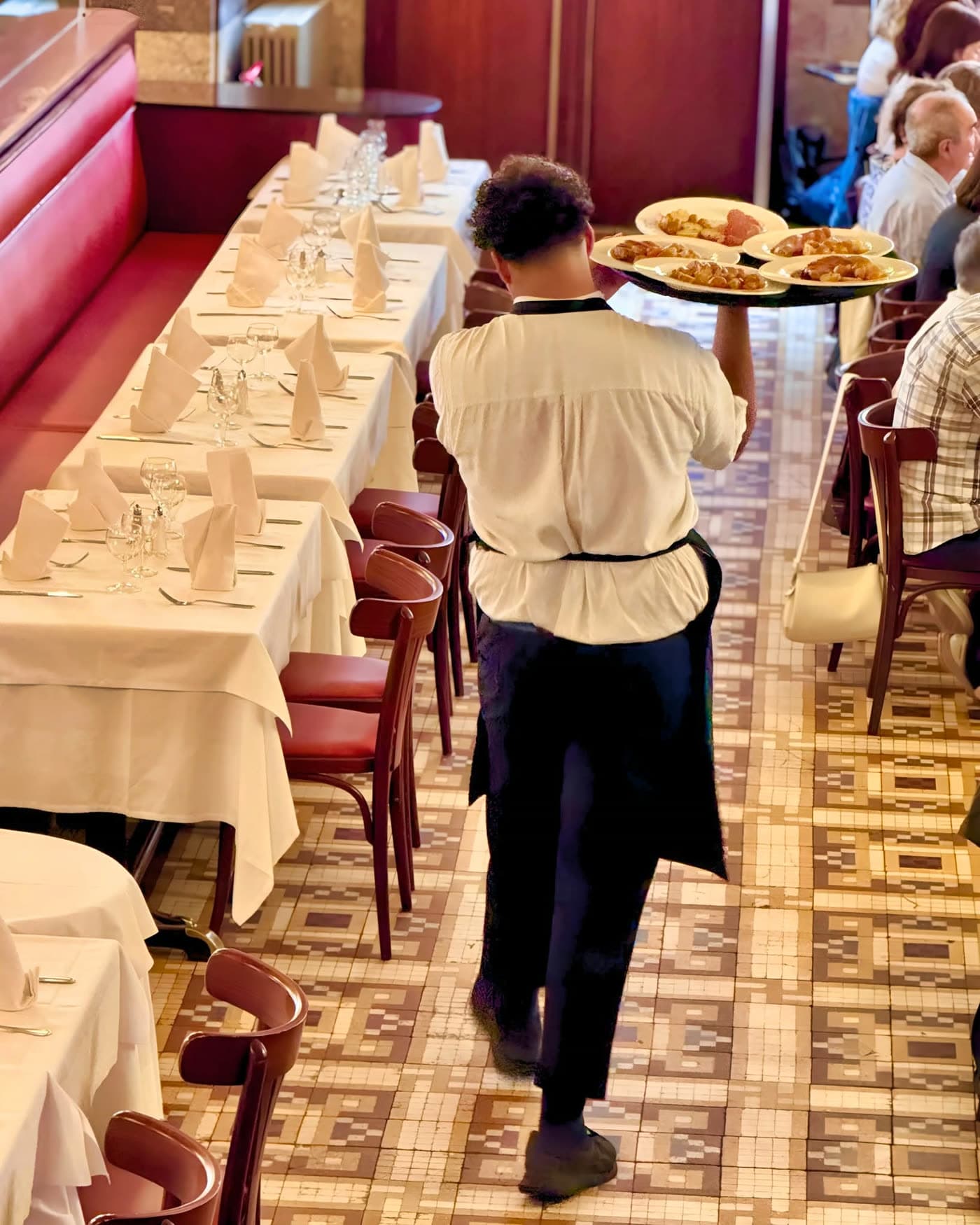 Serveur de dos portant un plateau de plats dans une allée entre tables dressées — Brasserie Georges