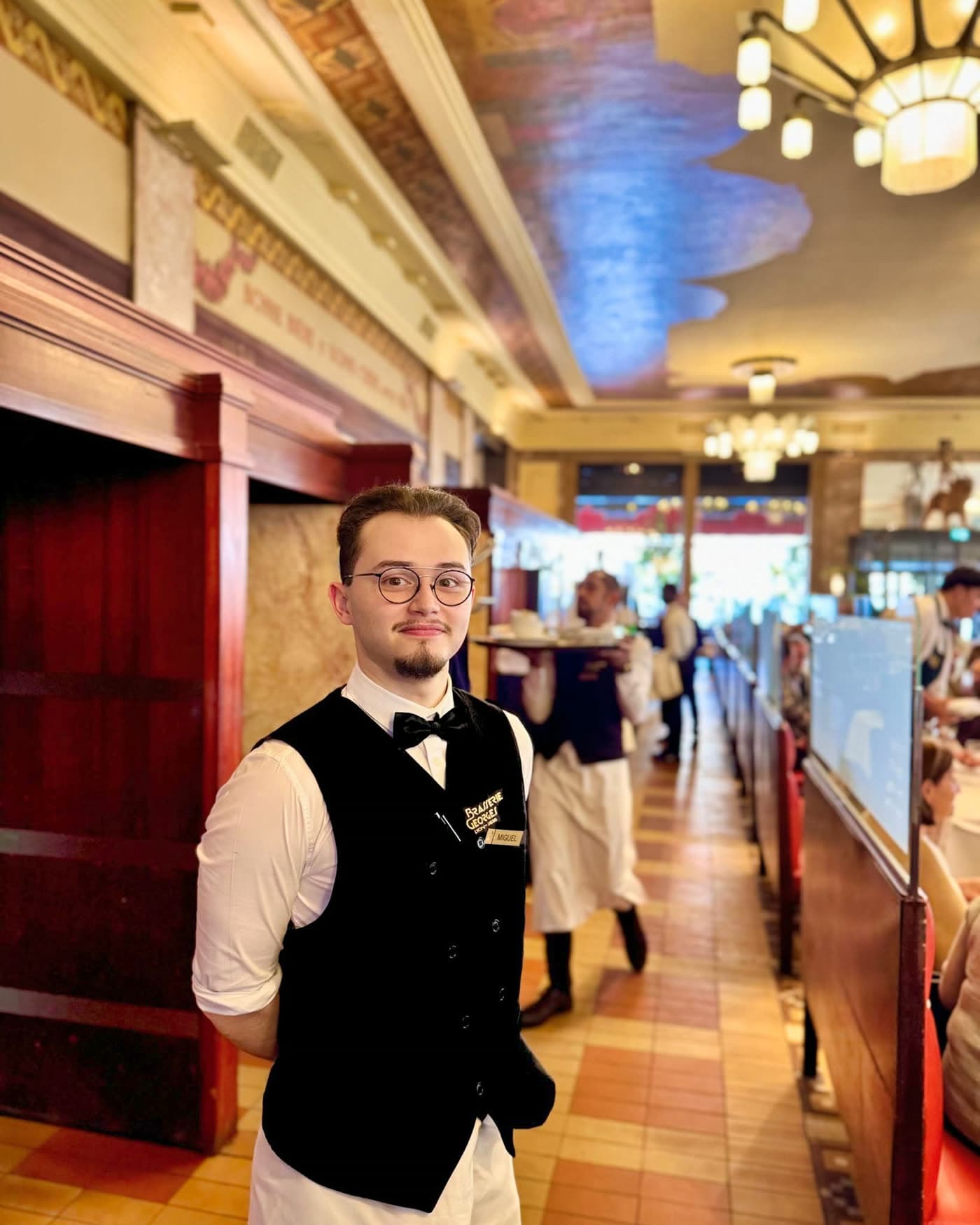Jeune serveur en gilet noir et noeud papillon souriant dans la salle, fresques au plafond en fond — Brasserie Georges