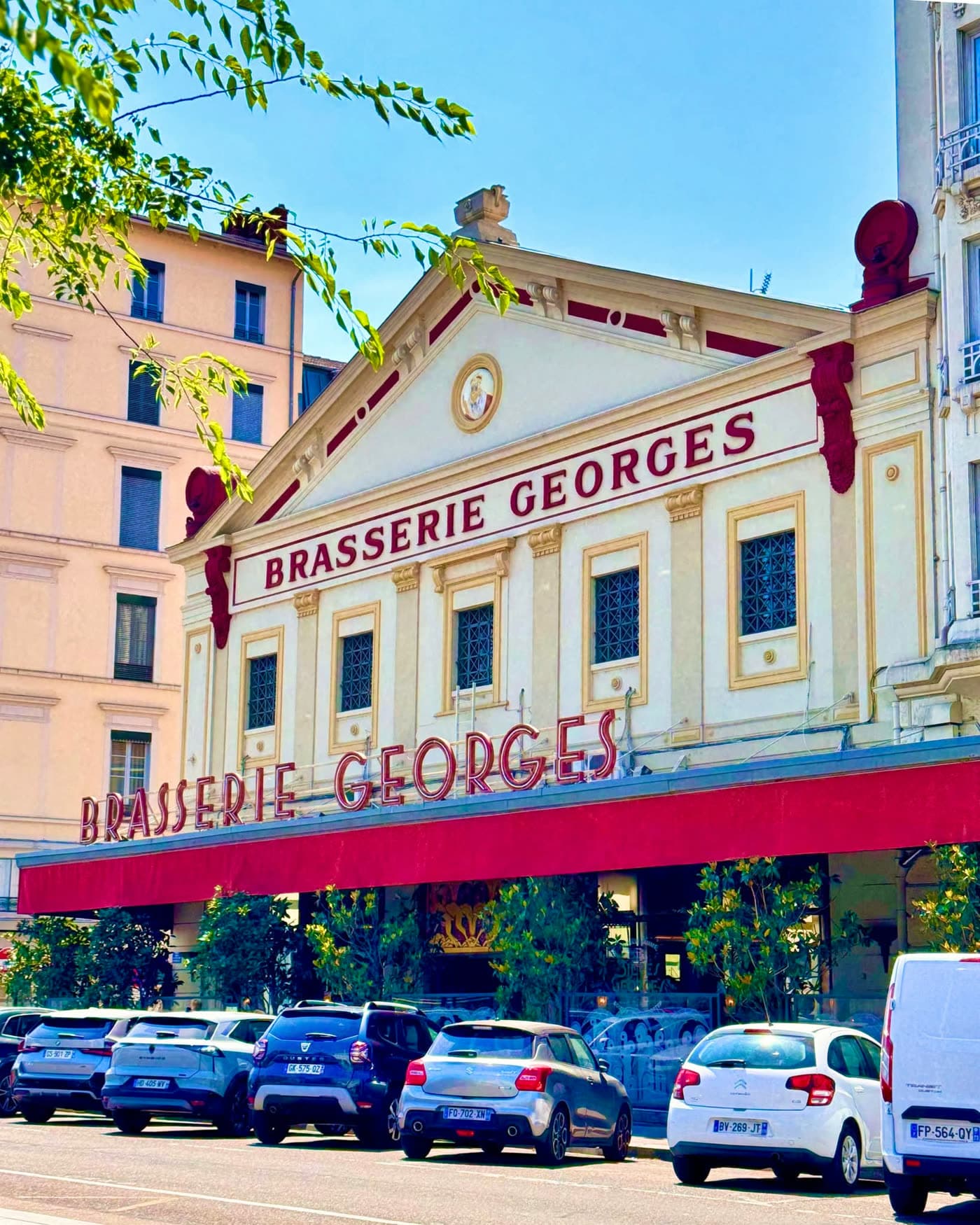 Façade de la Brasserie Georges de jour, enseigne rouge, ciel bleu, voitures garées devant