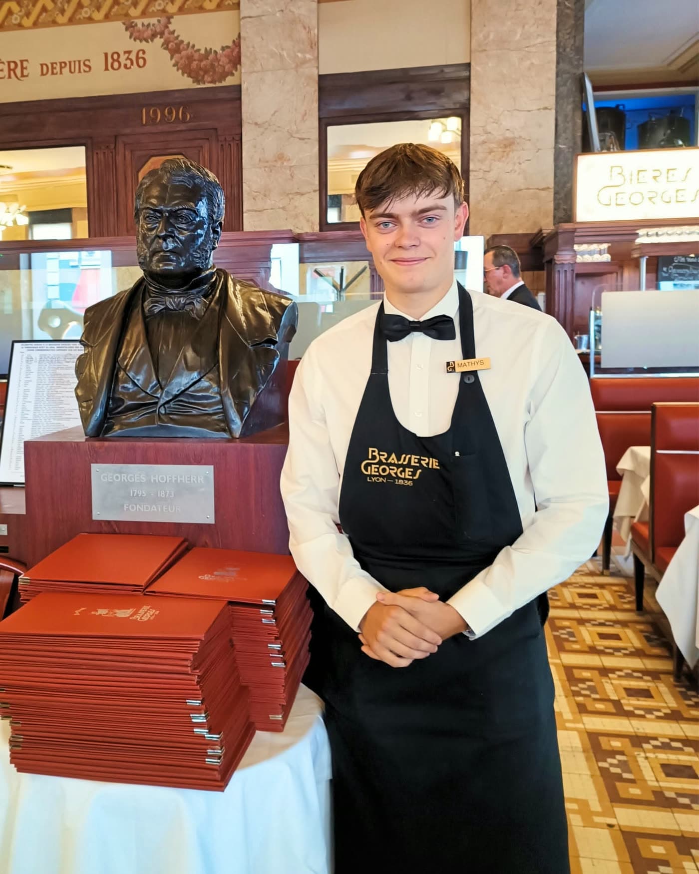 Serveur souriant posant près du buste du fondateur et d'une pile de menus rouges — Brasserie Georges