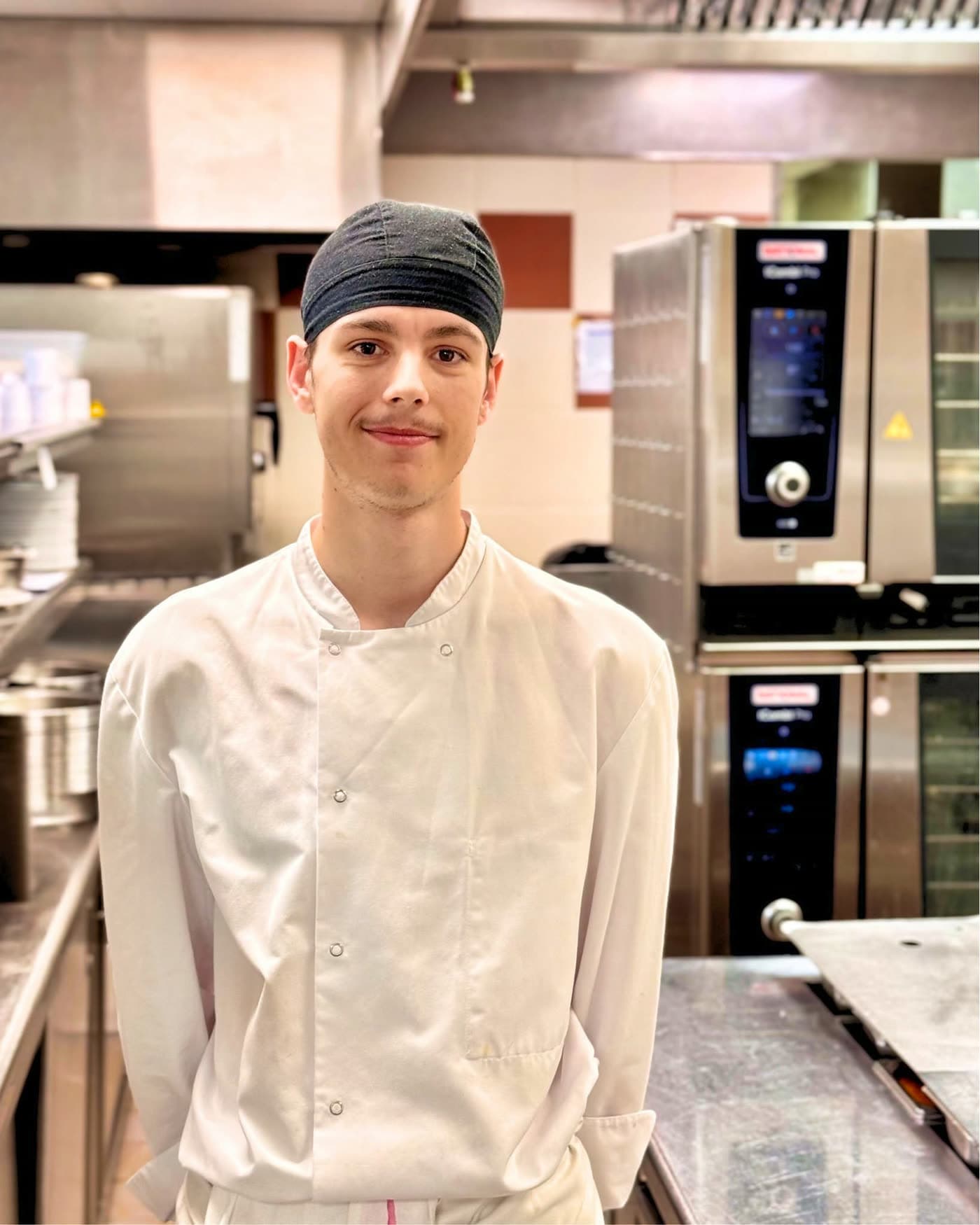 Jeune cuisinier en toque et veste blanche posant en cuisine devant des fours professionnels — Brasserie Georges