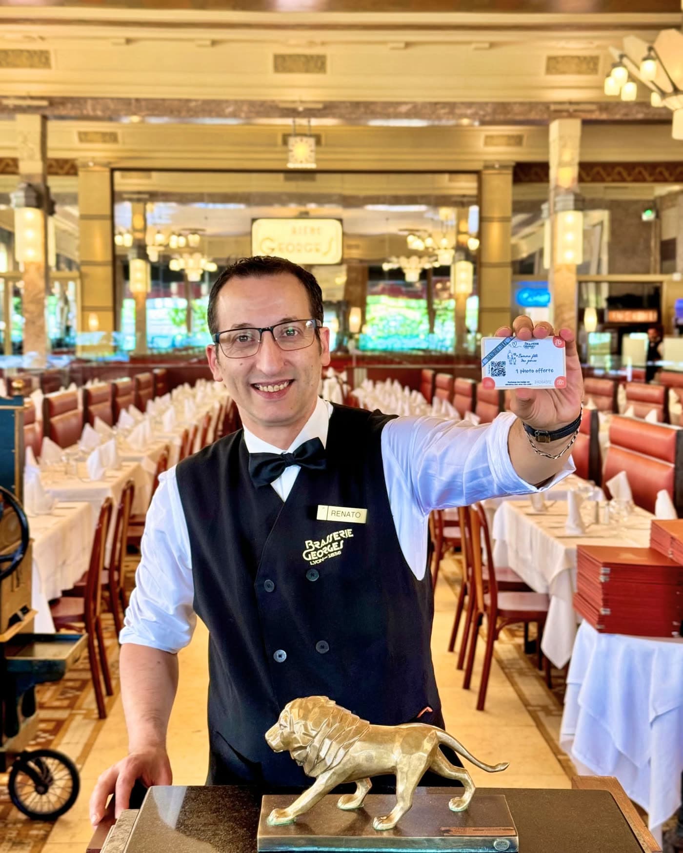 Serveur souriant tenant une carte de visite, statue de lion doré au premier plan, salle en fond — Brasserie Georges