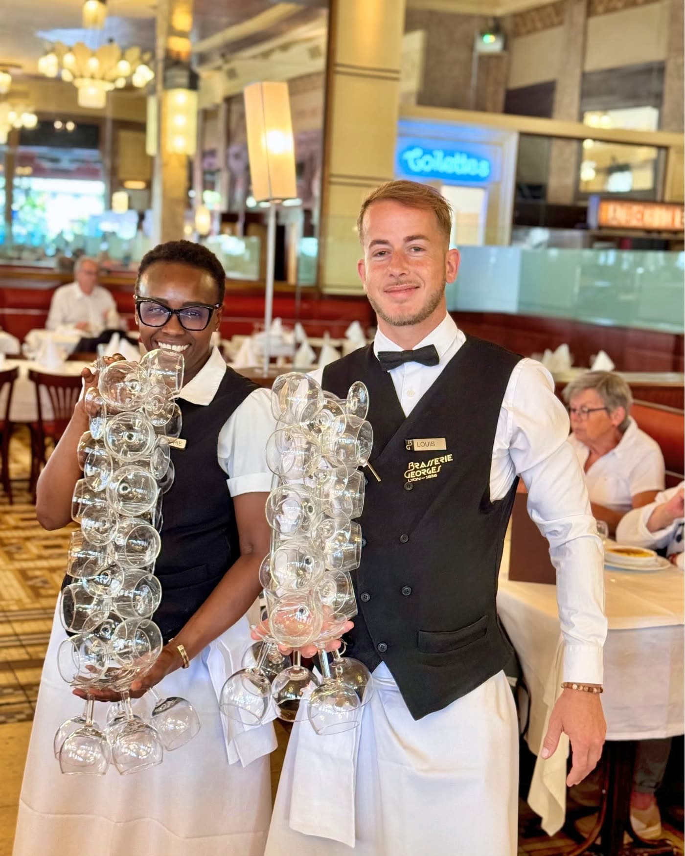 Deux serveurs portant chacun une longue pile de verres à vin empilés dans la salle — Brasserie Georges