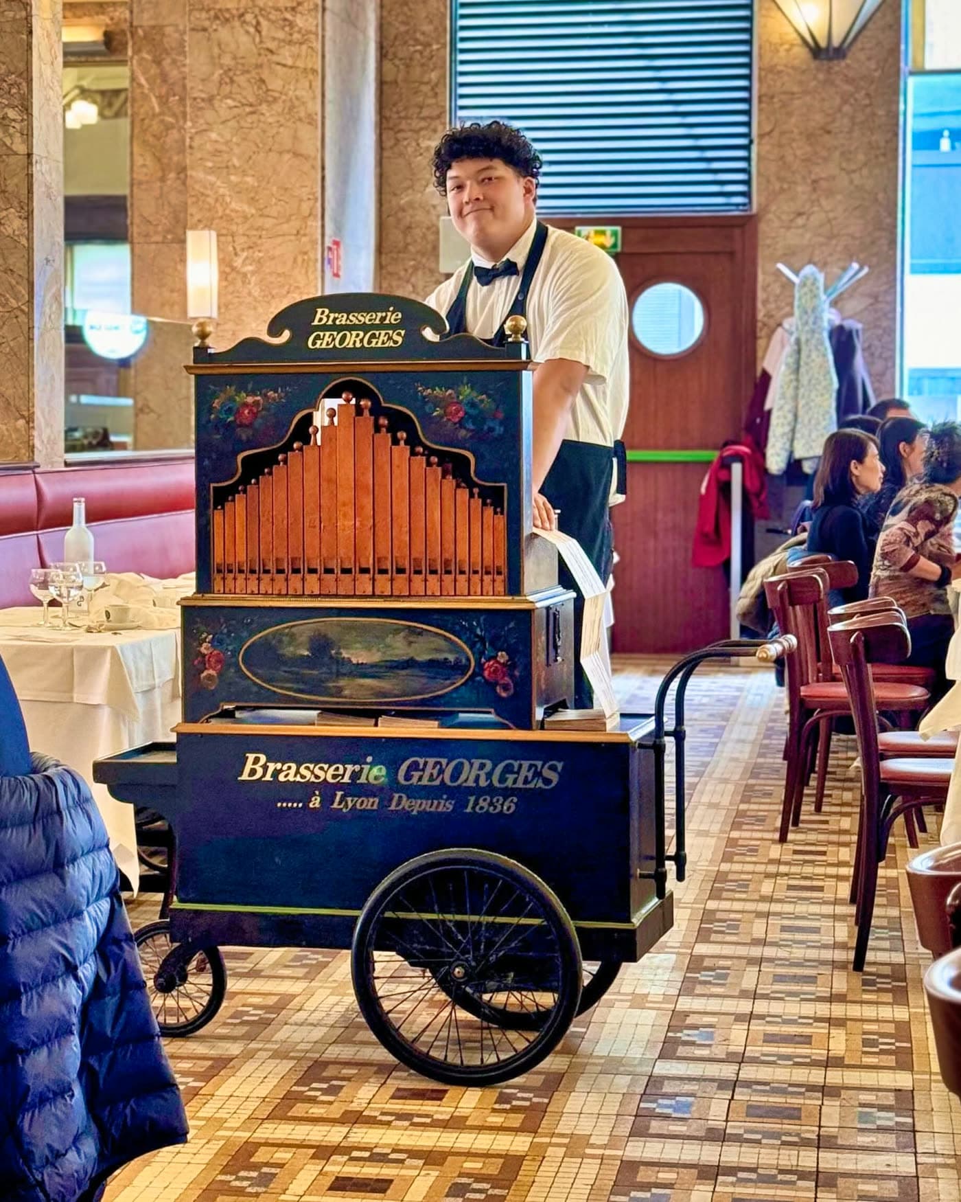 Jeune serveur souriant à côté d'un orgue de barbarie sur chariot Brasserie Georges dans la salle