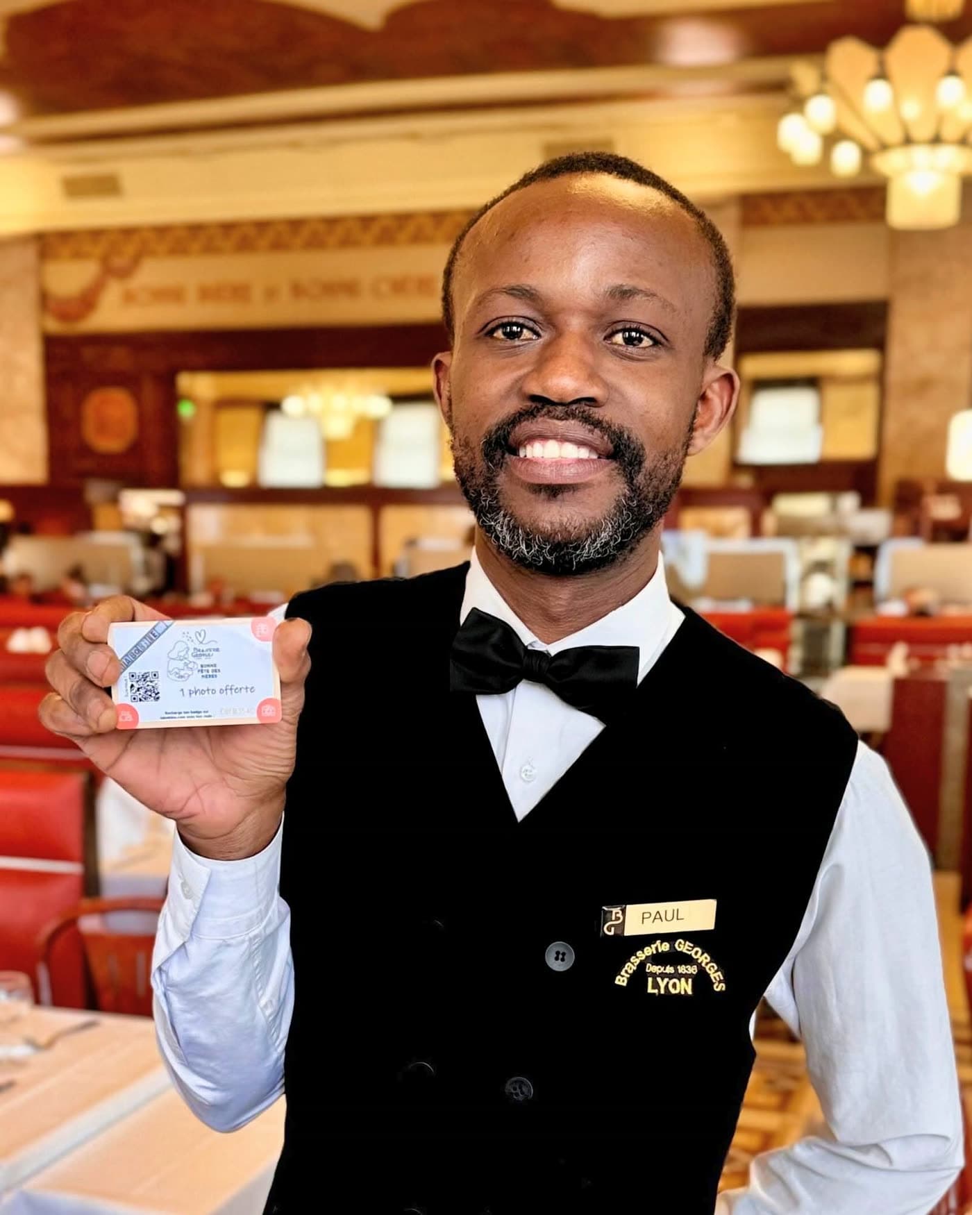 Serveur souriant en gilet noir tenant une carte de visite, salle et lustres en arrière-plan — Brasserie Georges