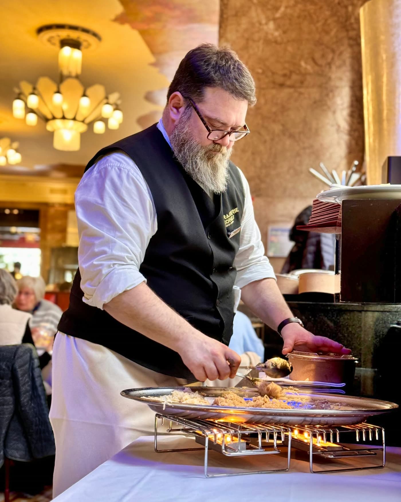 Maître d'hôtel barbu flambant un plat au guéridon devant la clientèle, flammes visibles — Brasserie Georges