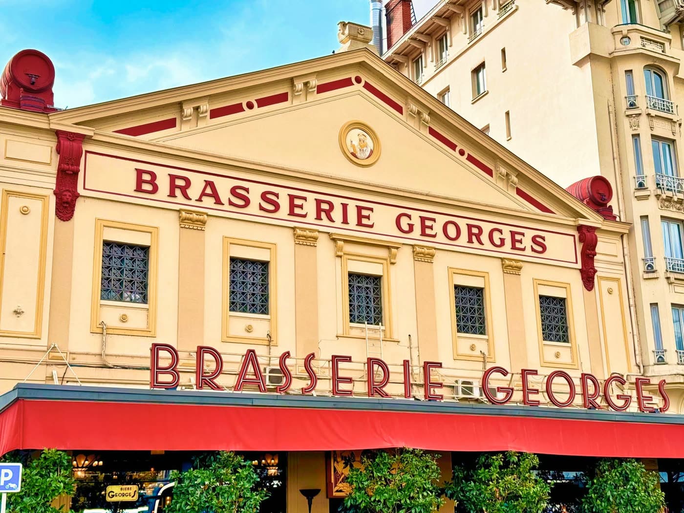 Façade Brasserie Georges de jour sous ciel bleu, enseignes rouges, store rouge déployé