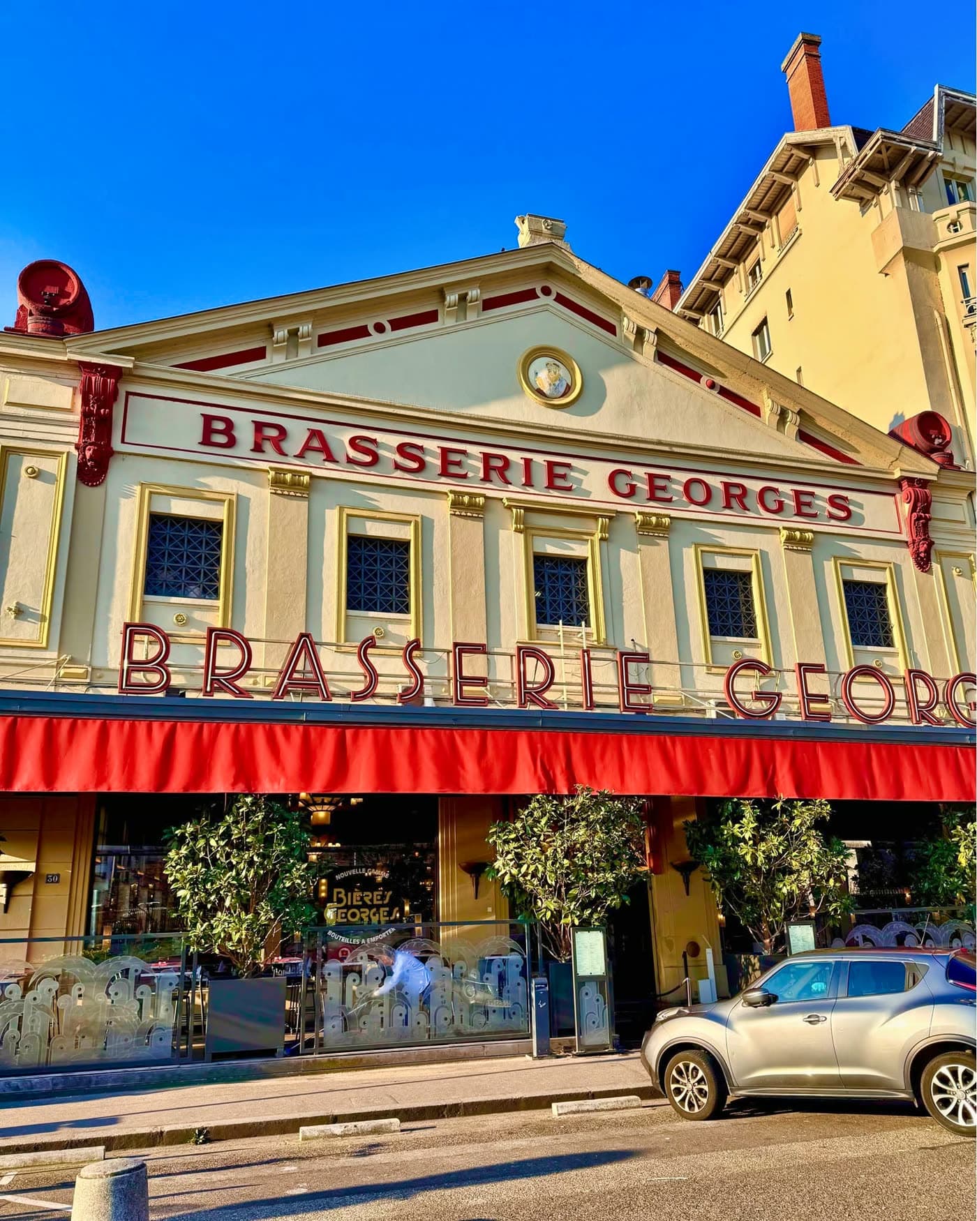 Façade Brasserie Georges de jour, store rouge déployé, ciel bleu, voiture garée devant