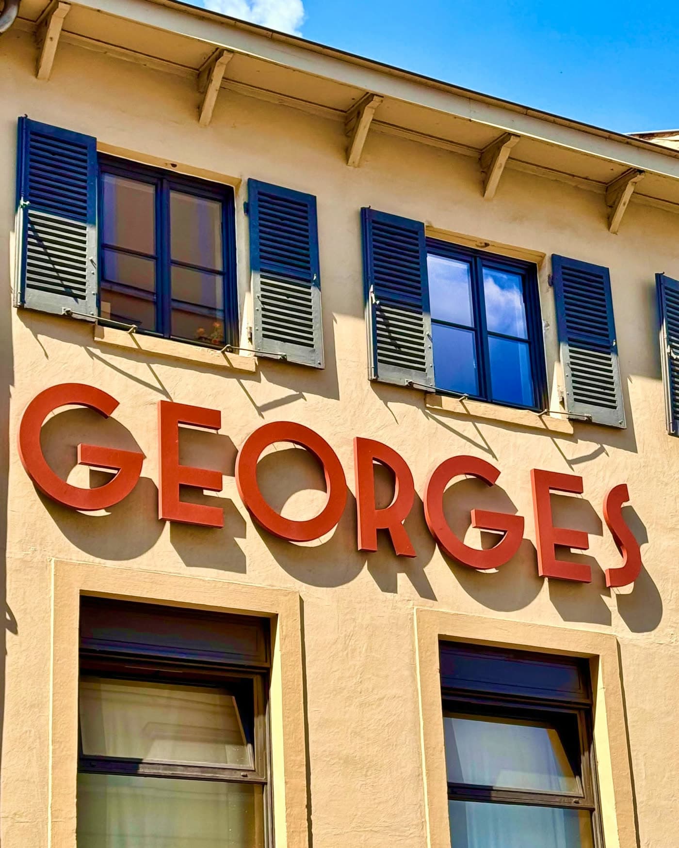 Gros plan sur l'enseigne rouge GEORGES en façade avec volets bleus et fenêtres à l'étage — Brasserie Georges