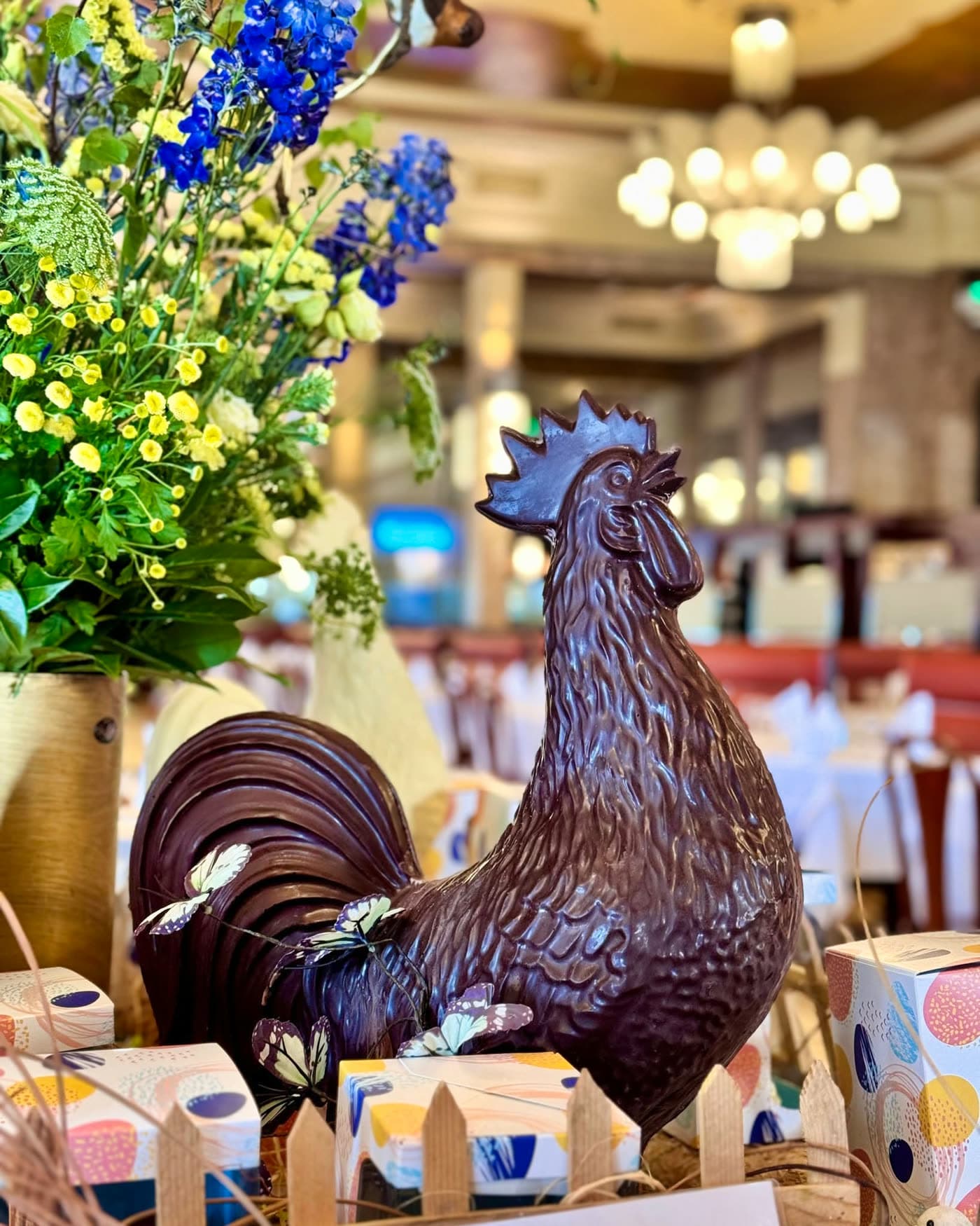 Coq en chocolat noir orné de papillons décoratifs, bouquet et lustre en fond — Brasserie Georges