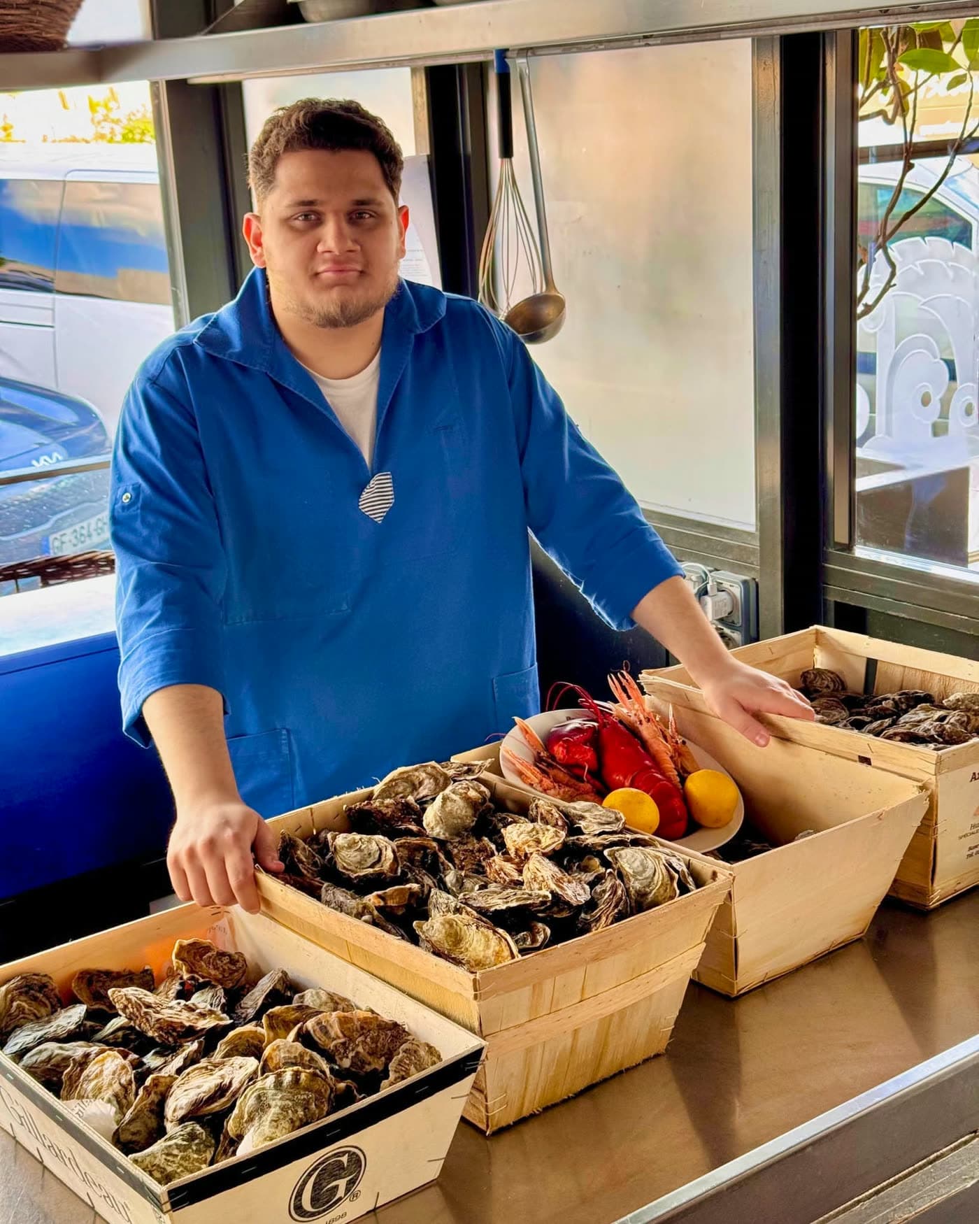 Jeune cuisinier posant devant des caisses d'huîtres, langoustines et citrons — Brasserie Georges
