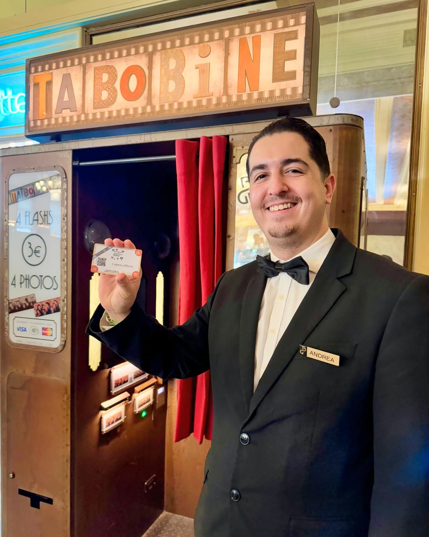 Serveur souriant tenant un ticket devant une cabine photomaton rétro 'Tabobine' — Brasserie Georges
