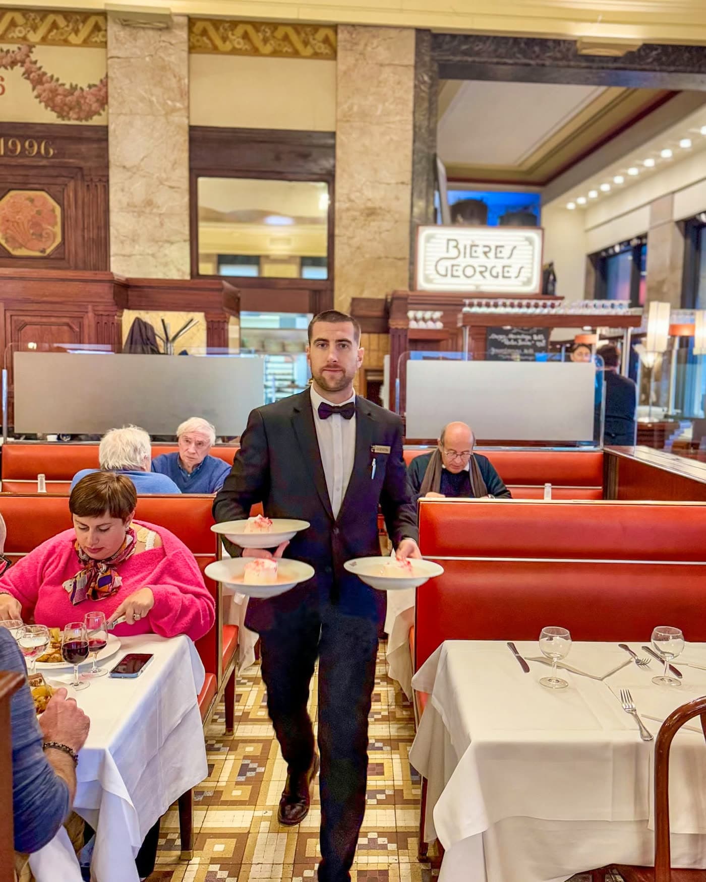 Serveur en nœud papillon portant trois assiettes empilées dans la salle sous l'enseigne Bières Georges — Brasserie Georges