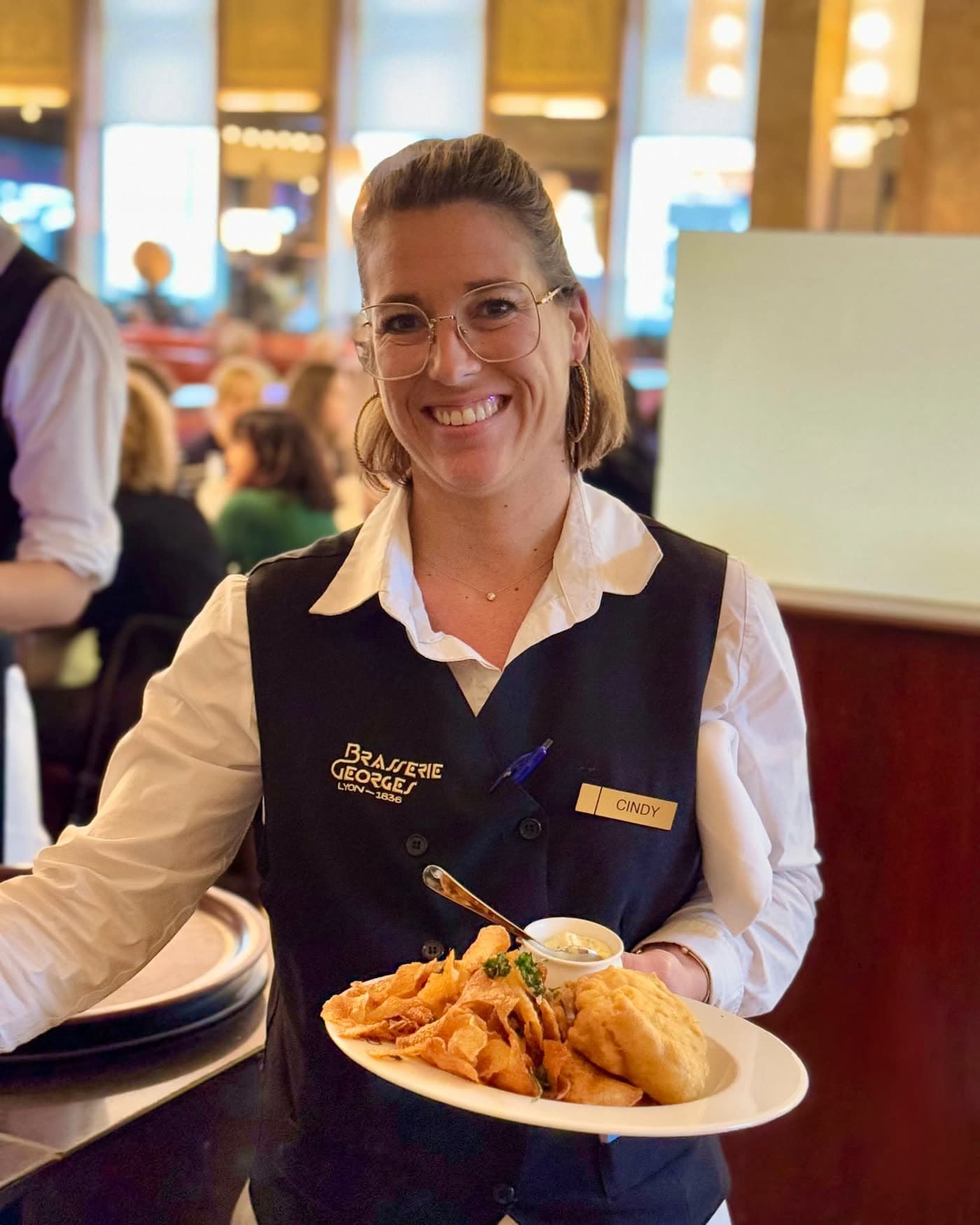 Serveuse en gilet Brasserie Georges présentant une assiette de fish and chips