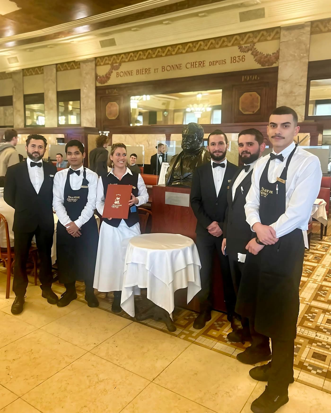 Six serveurs en tenue posant devant le buste sous fresque historique 1836 — Brasserie Georges