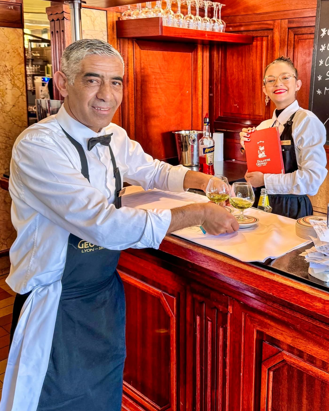 Barman servant des verres au comptoir en bois avec collègue tenant un livret rouge — Brasserie Georges