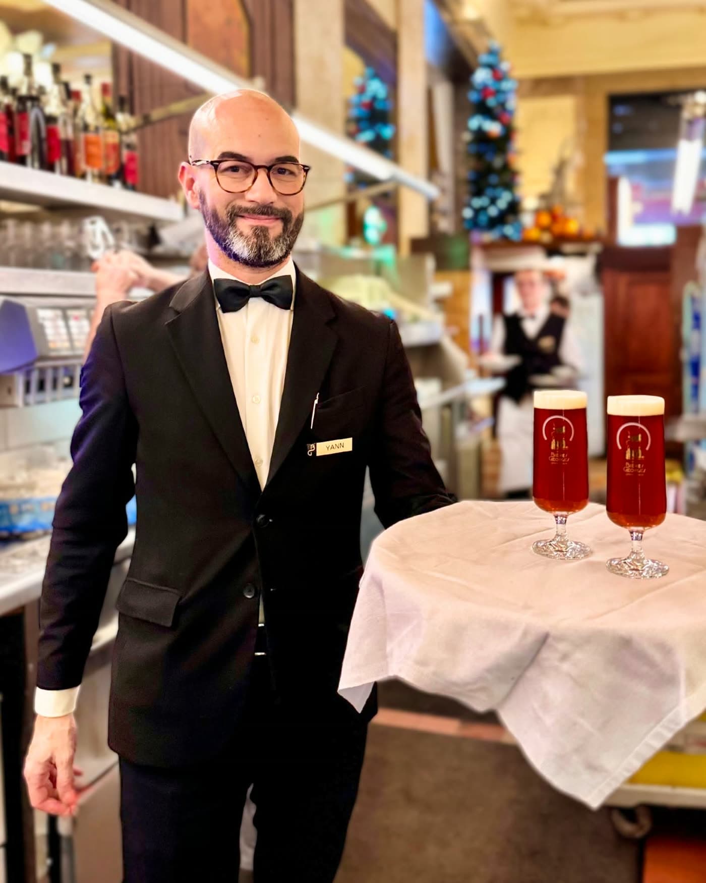 Serveur en smoking présentant deux verres de bière rousse sur plateau devant sapin de Noël — Brasserie Georges