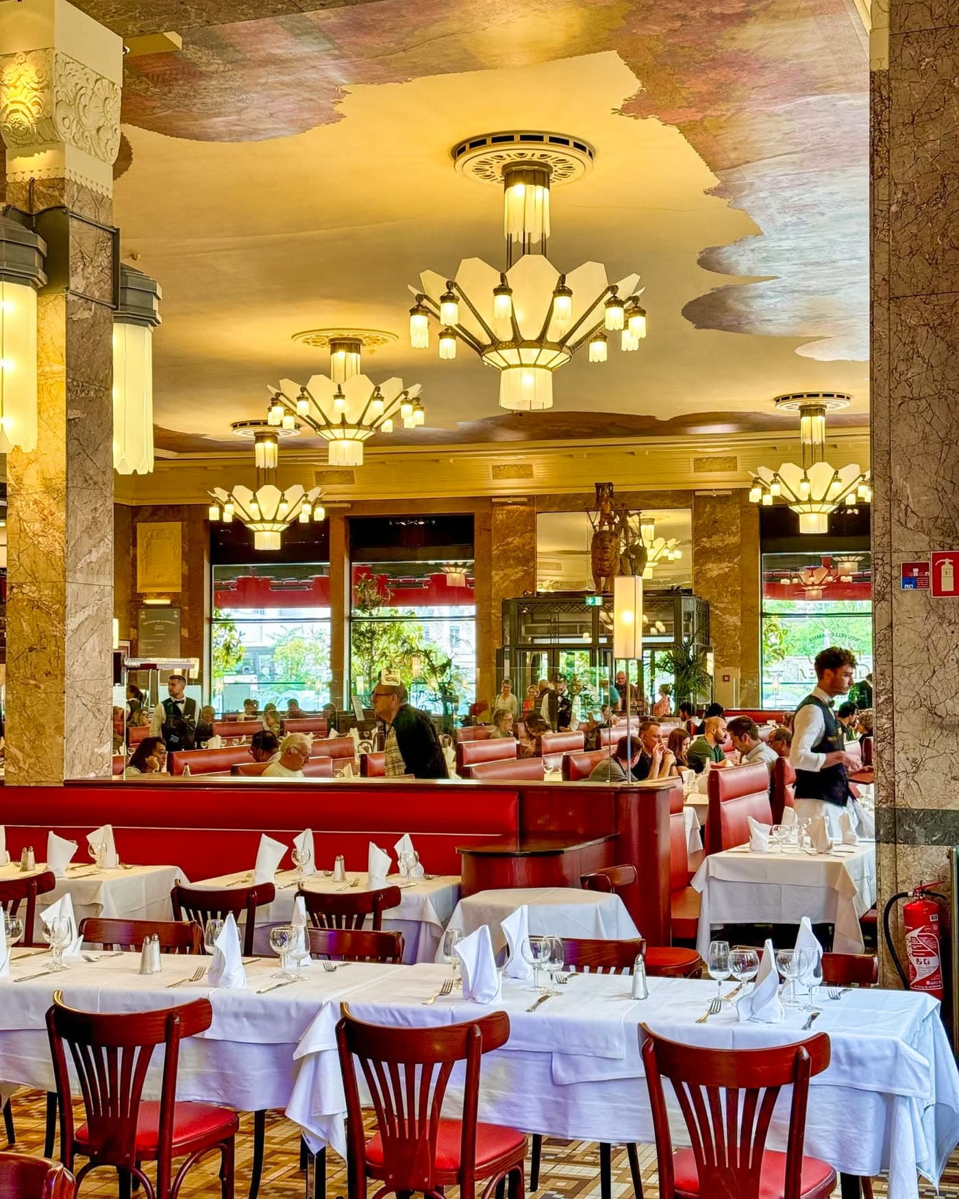 Salle Art déco vue large avec plafond peint, chandeliers, colonnes en marbre et tables dressées vides au premier plan — Brasserie Georges