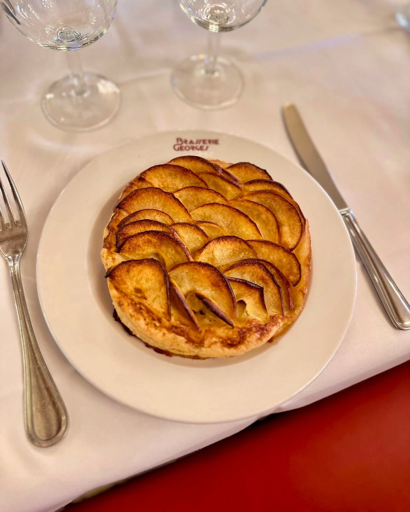 Tarte aux pommes dorée sur assiette blanche avec logo Brasserie Georges, couverts et verres à vin