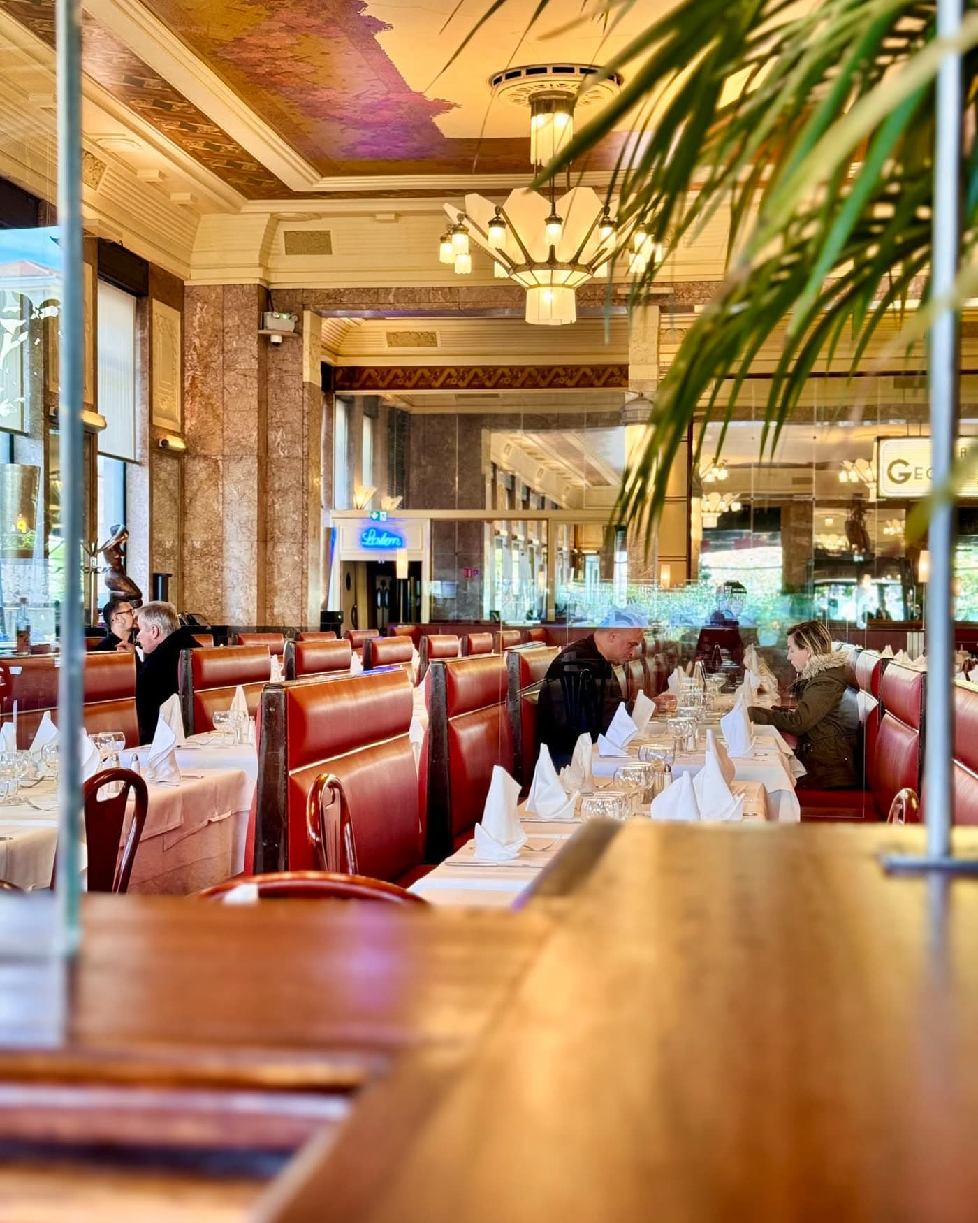 Vue de la salle avec feuilles de palmier au premier plan, banquettes rouges, chandeliers Art déco — Brasserie Georges