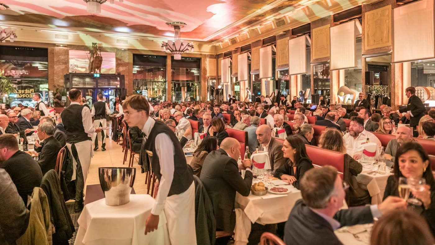 Vue large de la salle bondée en soirée avec serveur portant un seau à glace, chandeliers Art déco allumés — Brasserie Georges