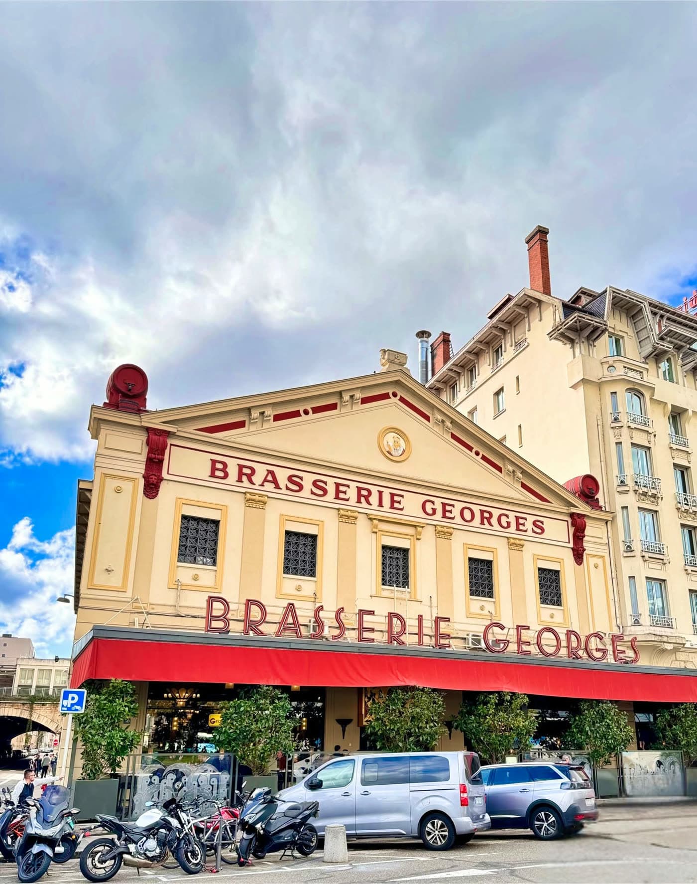 Façade complète de la Brasserie Georges de jour, auvent rouge, ciel nuageux, deux-roues garés