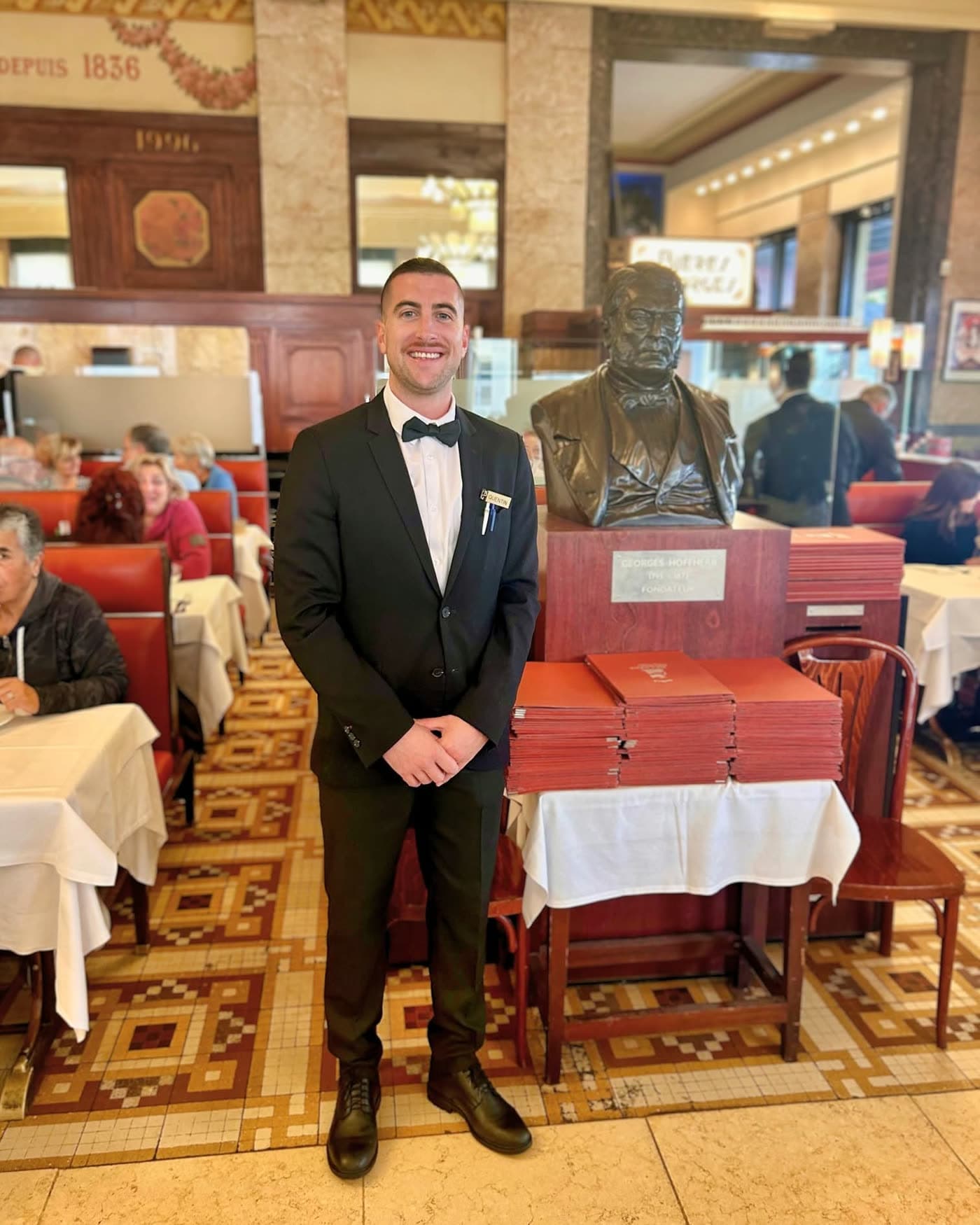 Serveur souriant en costume pose près d'un buste en bronze, salle en arrière-plan — Brasserie Georges
