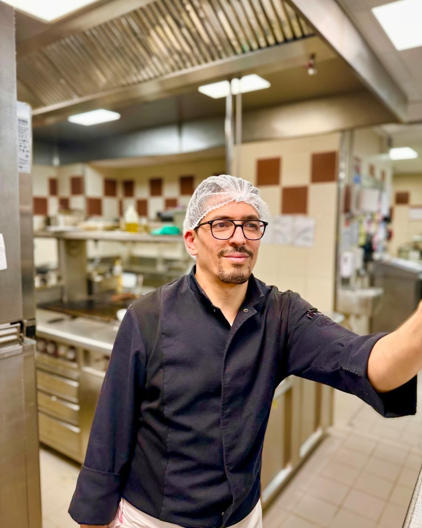 Chef en cuisine, charlotte et lunettes, sourit face à la caméra devant équipement inox — Brasserie Georges