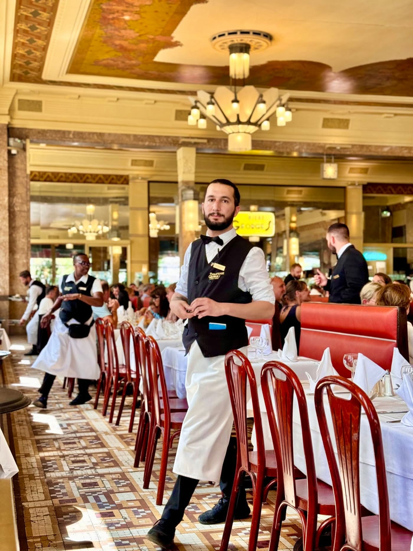 Serveur debout dans la salle, plafond peint Art déco et lustres visibles en arrière-plan — Brasserie Georges