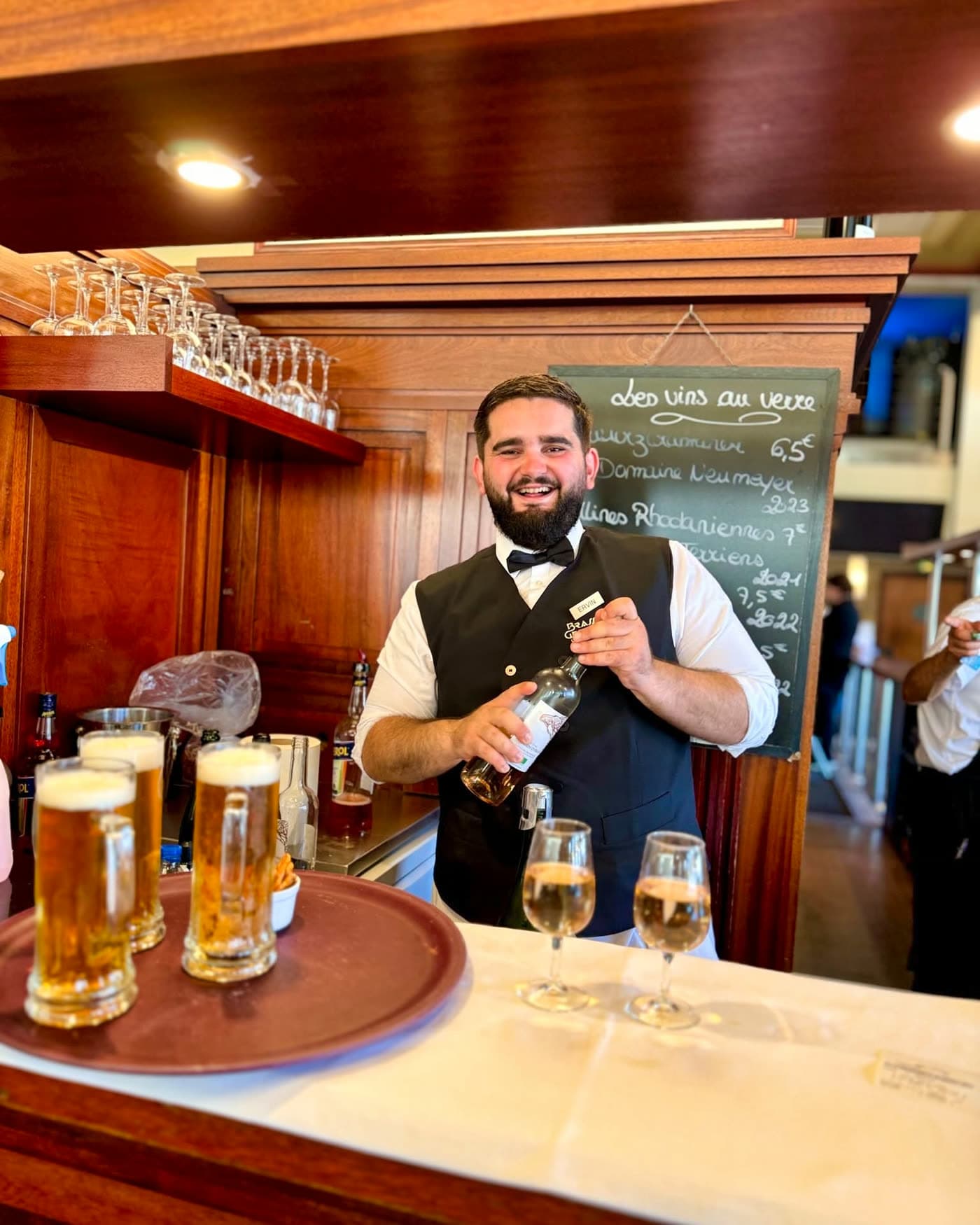Serveur souriant au bar présente une bouteille, verres de bière et flûtes de champagne — Brasserie Georges
