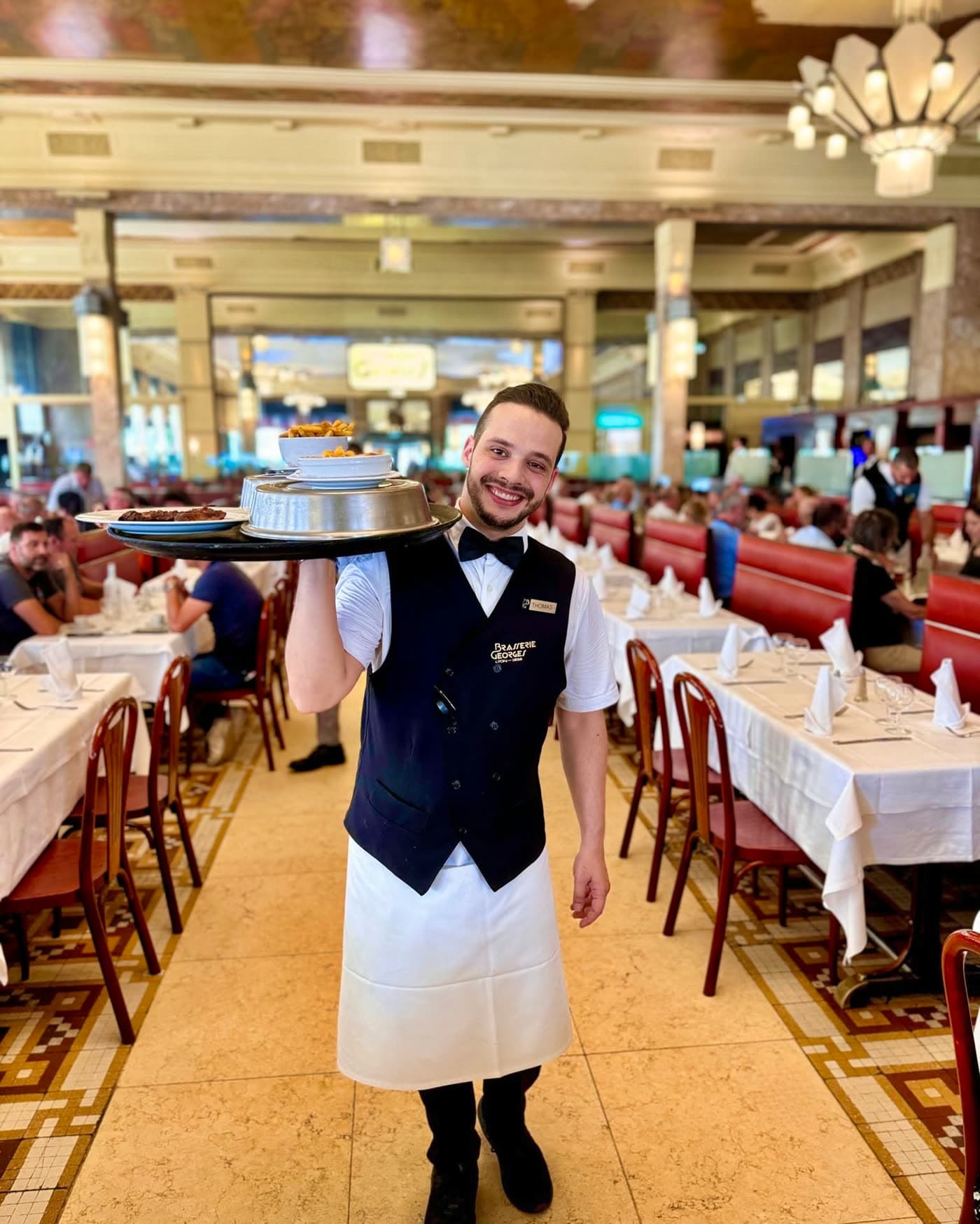 Serveur portant un plateau de cloches traverse la grande salle sous les lustres — Brasserie Georges