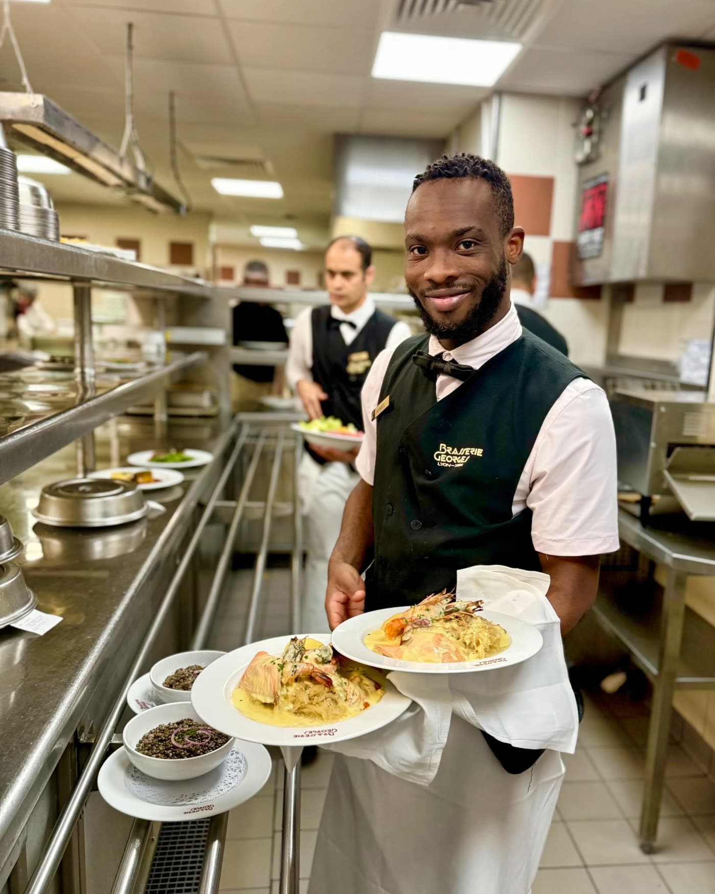 Serveur souriant portant plusieurs assiettes garnies dans le passe de cuisine — Brasserie Georges