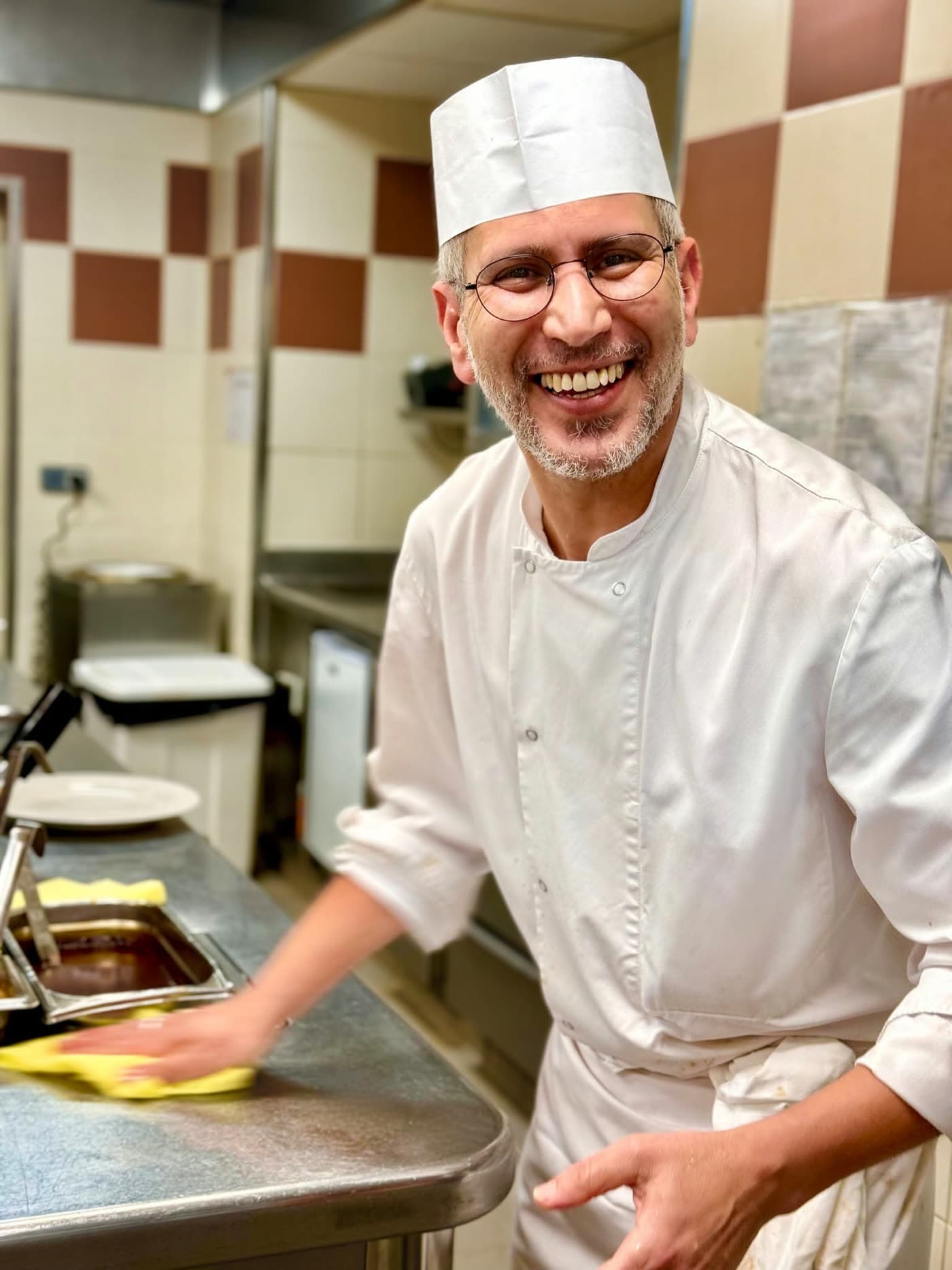 Chef en tenue blanche souriant largement en nettoyant un plan de travail inox — Brasserie Georges