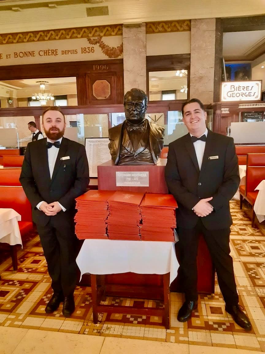 Deux serveurs en smoking posant près d'un buste en bronze et d'une pile de cartes rouges — Brasserie Georges
