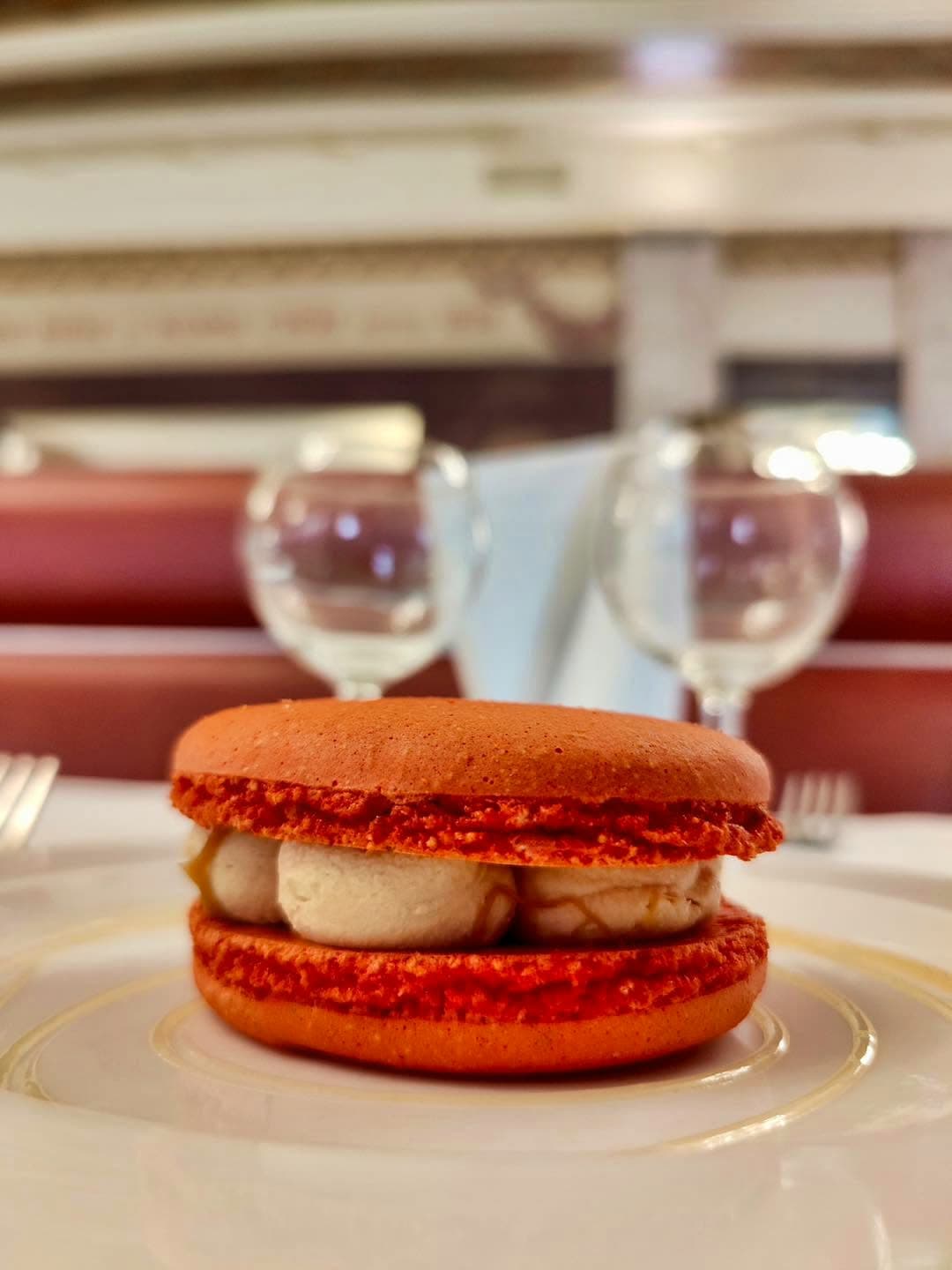 Gros plan sur un macaron rouge garni de crème, verres à vin flous en fond — Brasserie Georges
